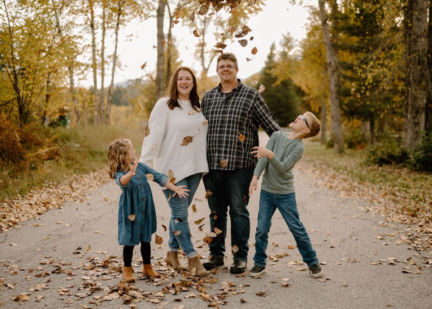 A joyful family of four tosses autumn leaves on a tree-lined path.
