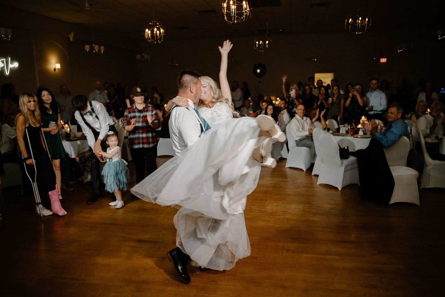 Groom lifts bride in flowing white dress during energetic first dance at elegant wedding reception.