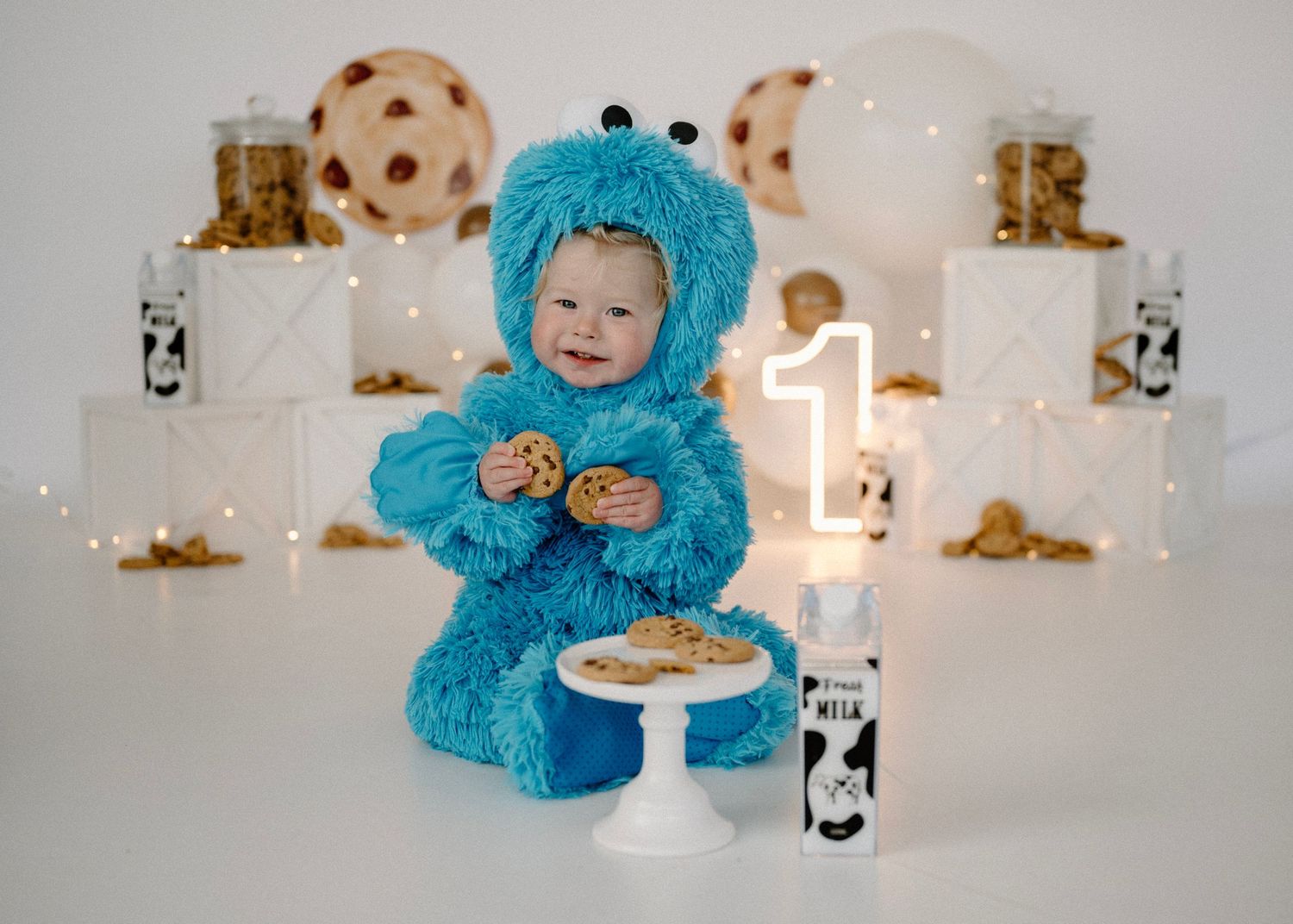 Baby dressed in Cookie Monster costume eating cookies beside number 1 candle and milk bottle.