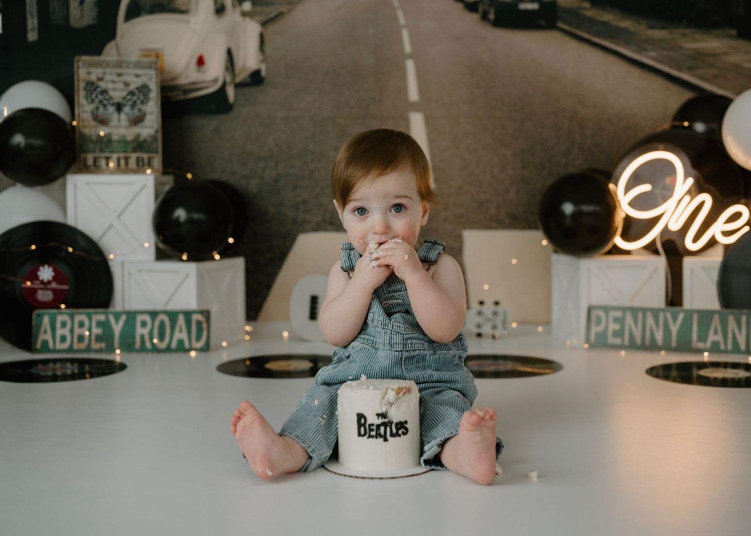 Toddler boy in denim overalls sits before Beatles-themed cake with Abbey Road birthday party decorations.
