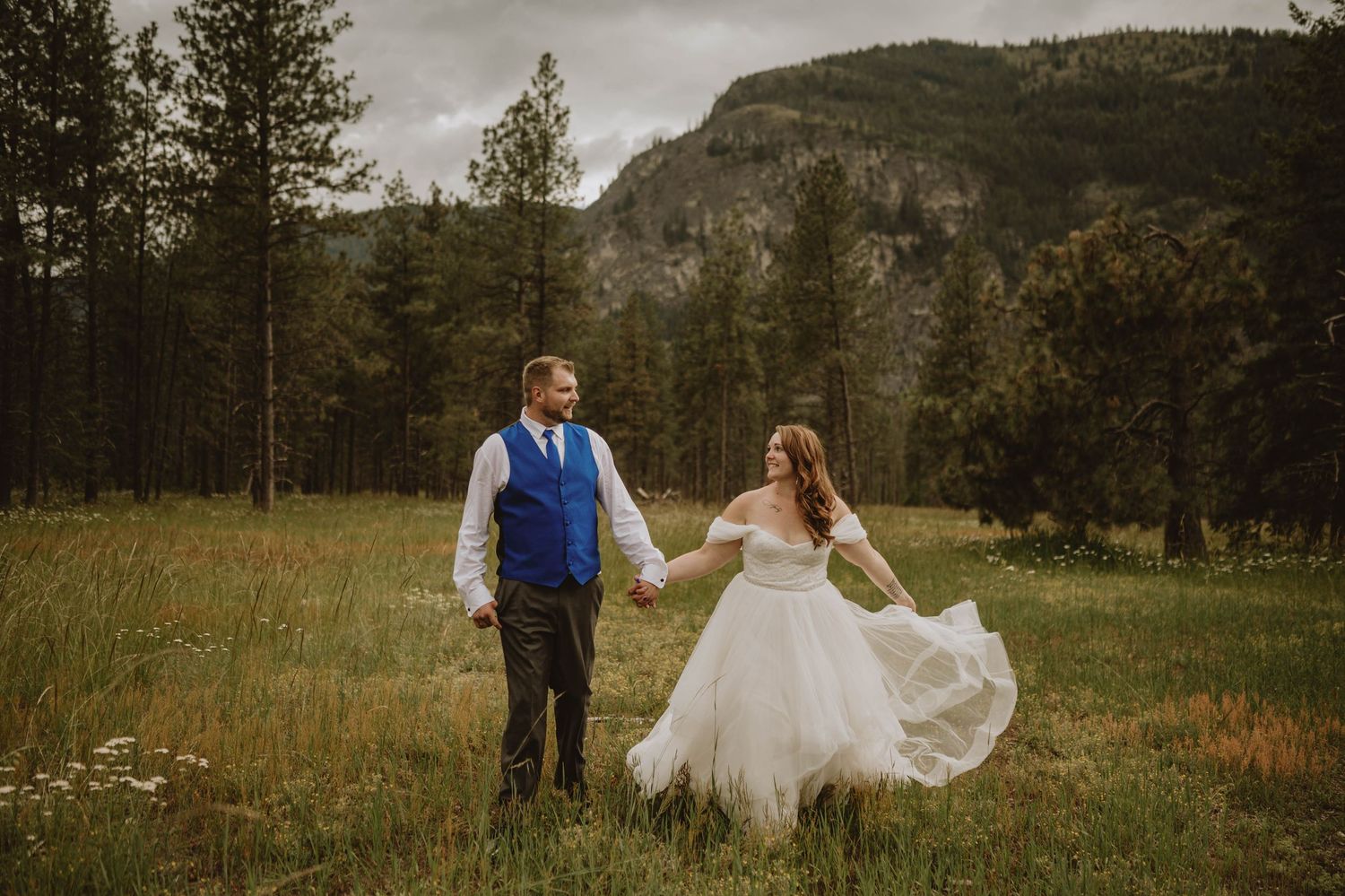 Bride and groom hold hands in mountain meadow, wedding dress flowing in the breeze.