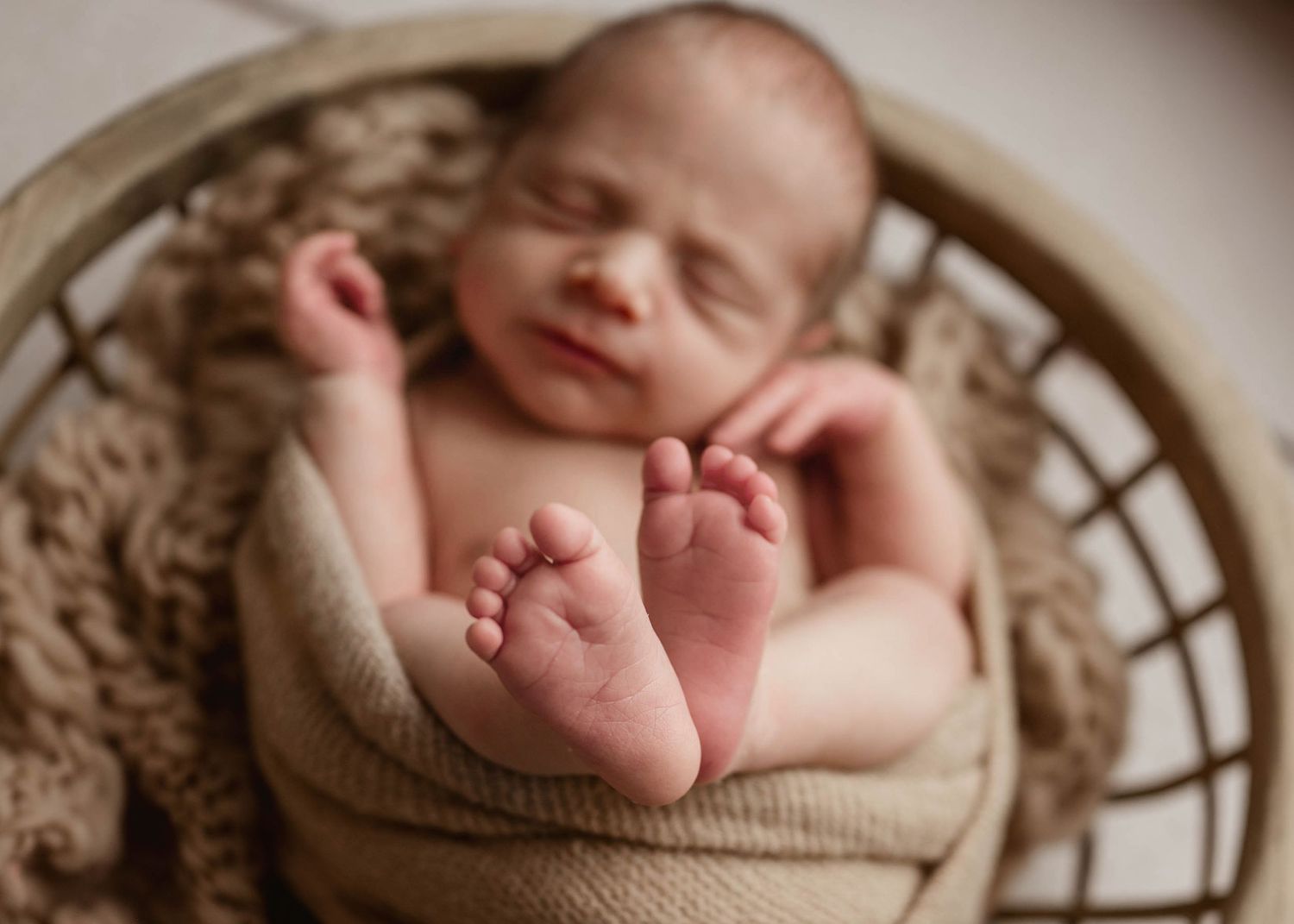 Sleeping smiling newborn baby wrapped in beige knit in round wicker basket with tiny feet visible.