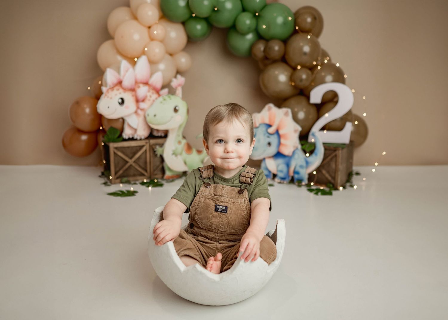 Happy toddler boy sits inside a white egg prop at a dinosaur-themed 2nd birthday cake smash session.