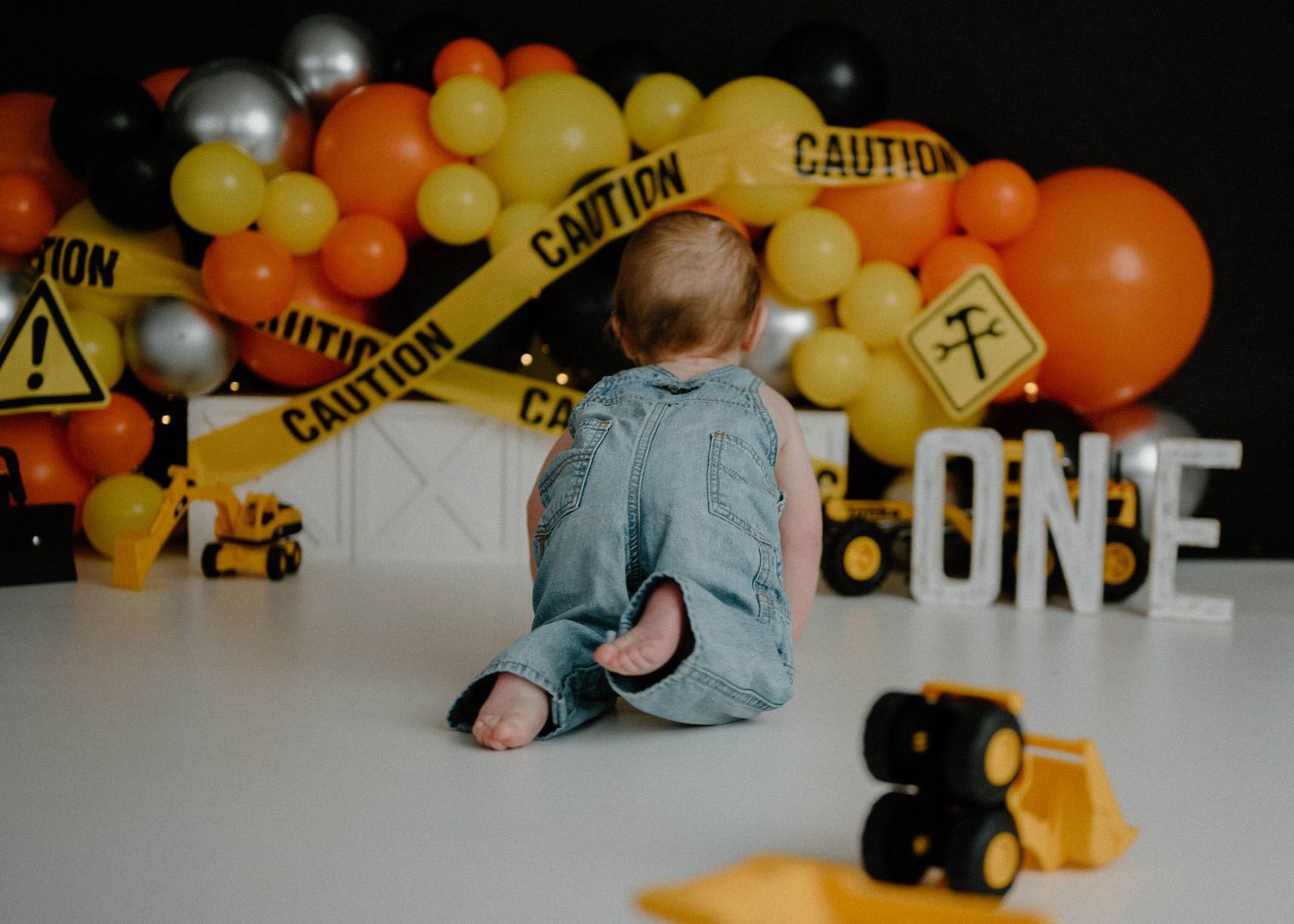 Baby in denim overalls sits facing construction-themed first birthday backdrop with orange and black balloons.