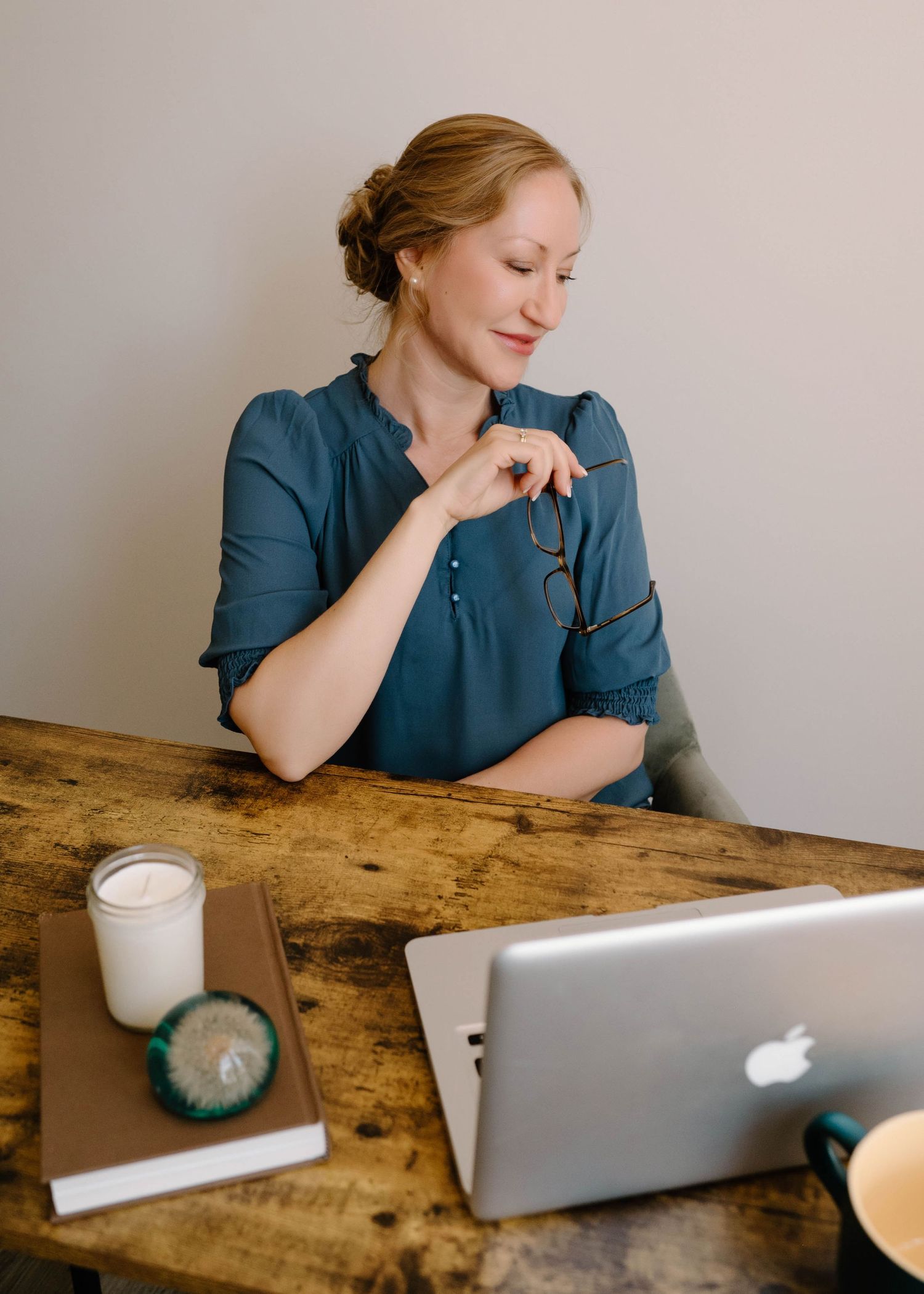 Blonde woman in teal blouse holding glasses, sitting at rustic wooden desk with MacBook laptop.