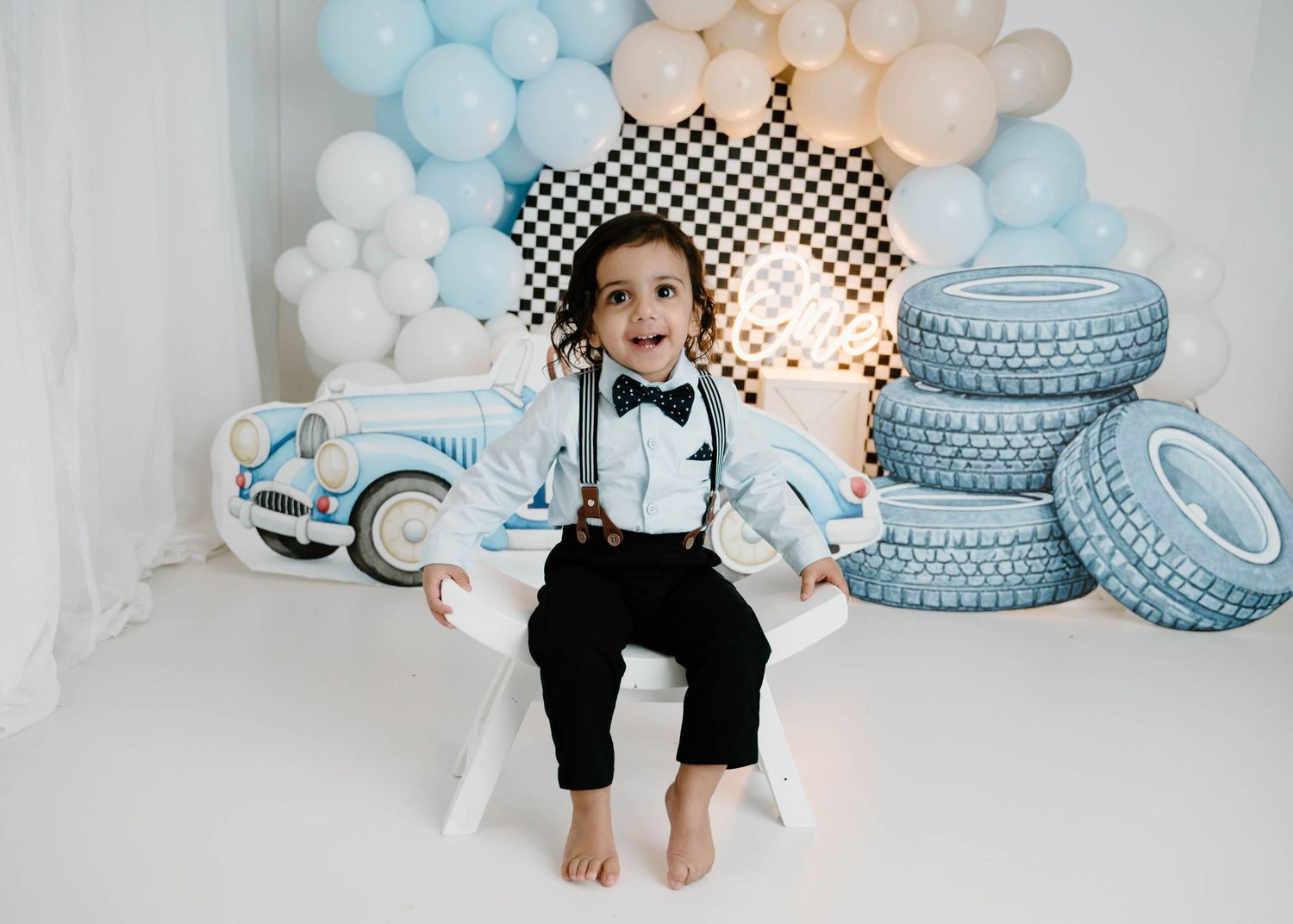 A smiling toddler boy in suspenders and bow tie sits on a white bench at a vintage race car first birthday photo shoot.