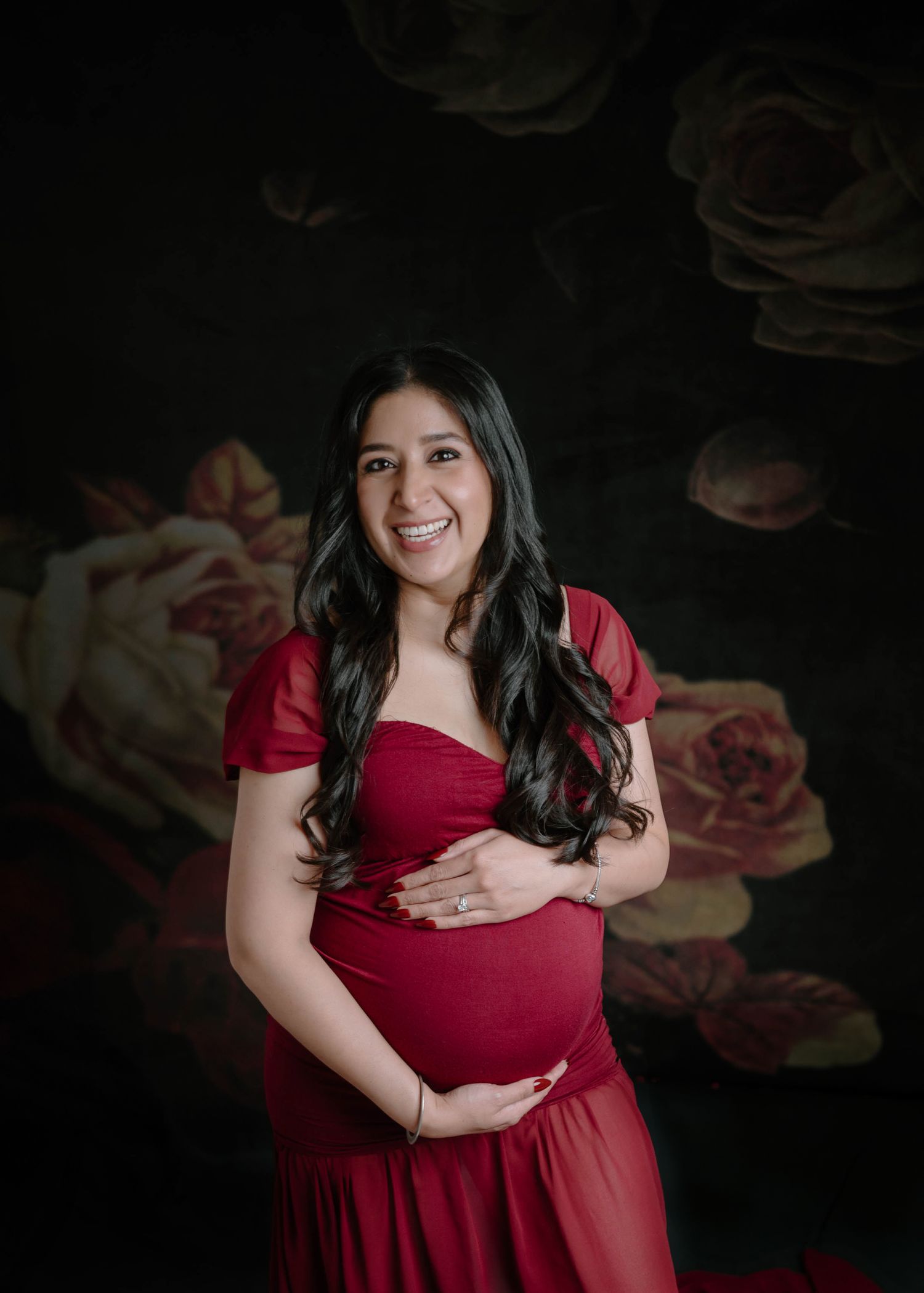 A smiling pregnant woman in a red dress cradles her bump against a dark floral background.