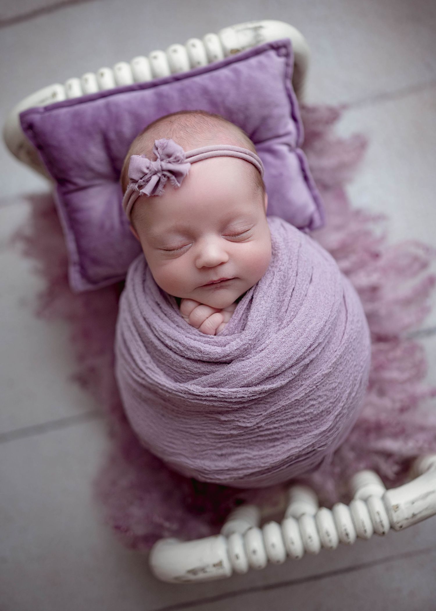Sleeping newborn baby swaddled in purple wrap with bow headband on tiny white bed with lavender pillow and fluffy rug.