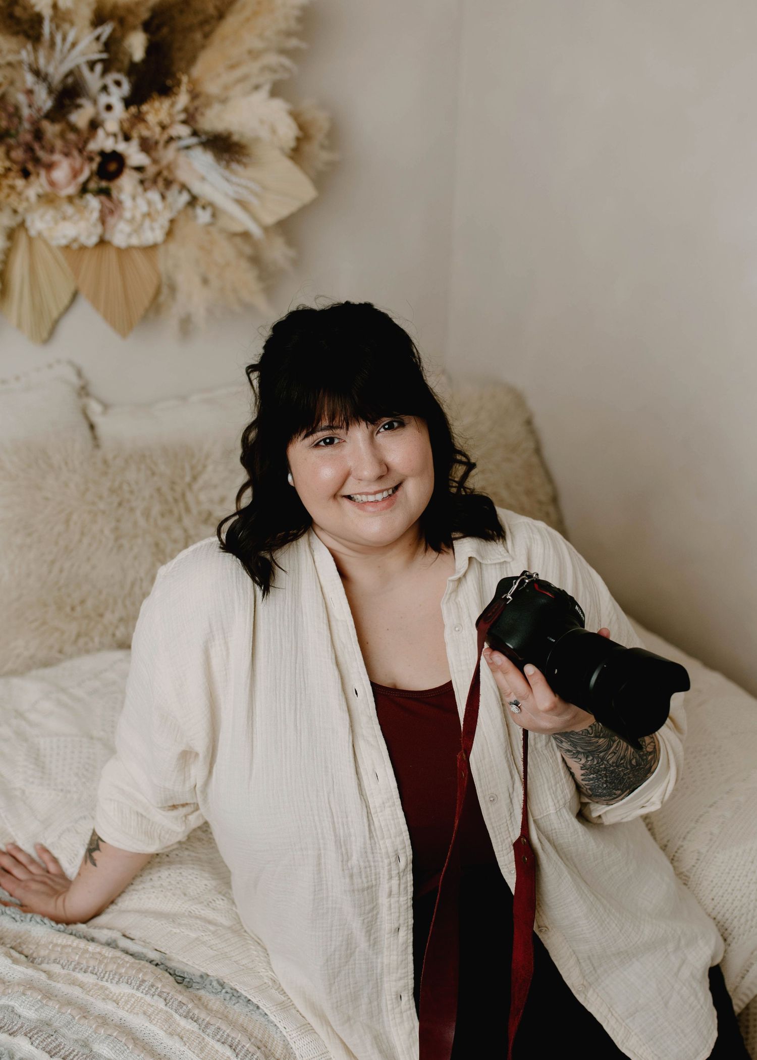 Female photographer smiling and holding camera while sitting on couch with boho floral wall decor.