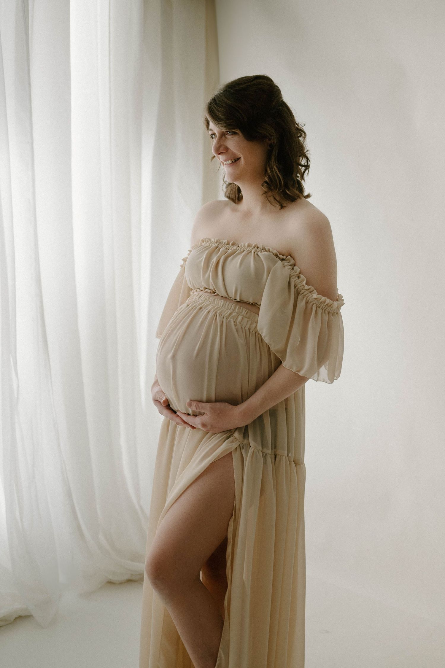 Smiling pregnant woman in elegant off-shoulder beige chiffon gown cradles belly in studio.