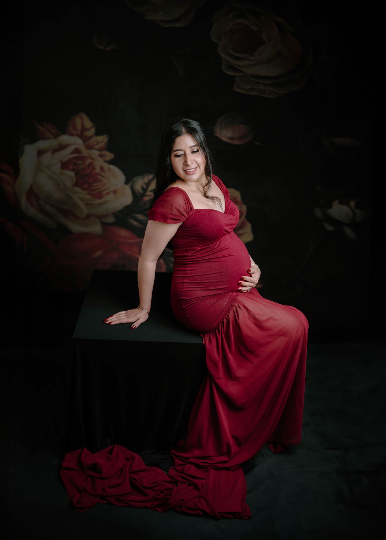 Smiling pregnant woman in red maternity gown posed on black cube with floral background.