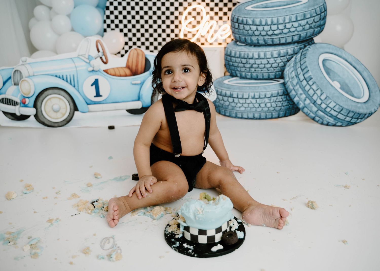 A baby boy in a black romper sits on the floor doing a cake smash at a vintage race car themed first birthday shoot.