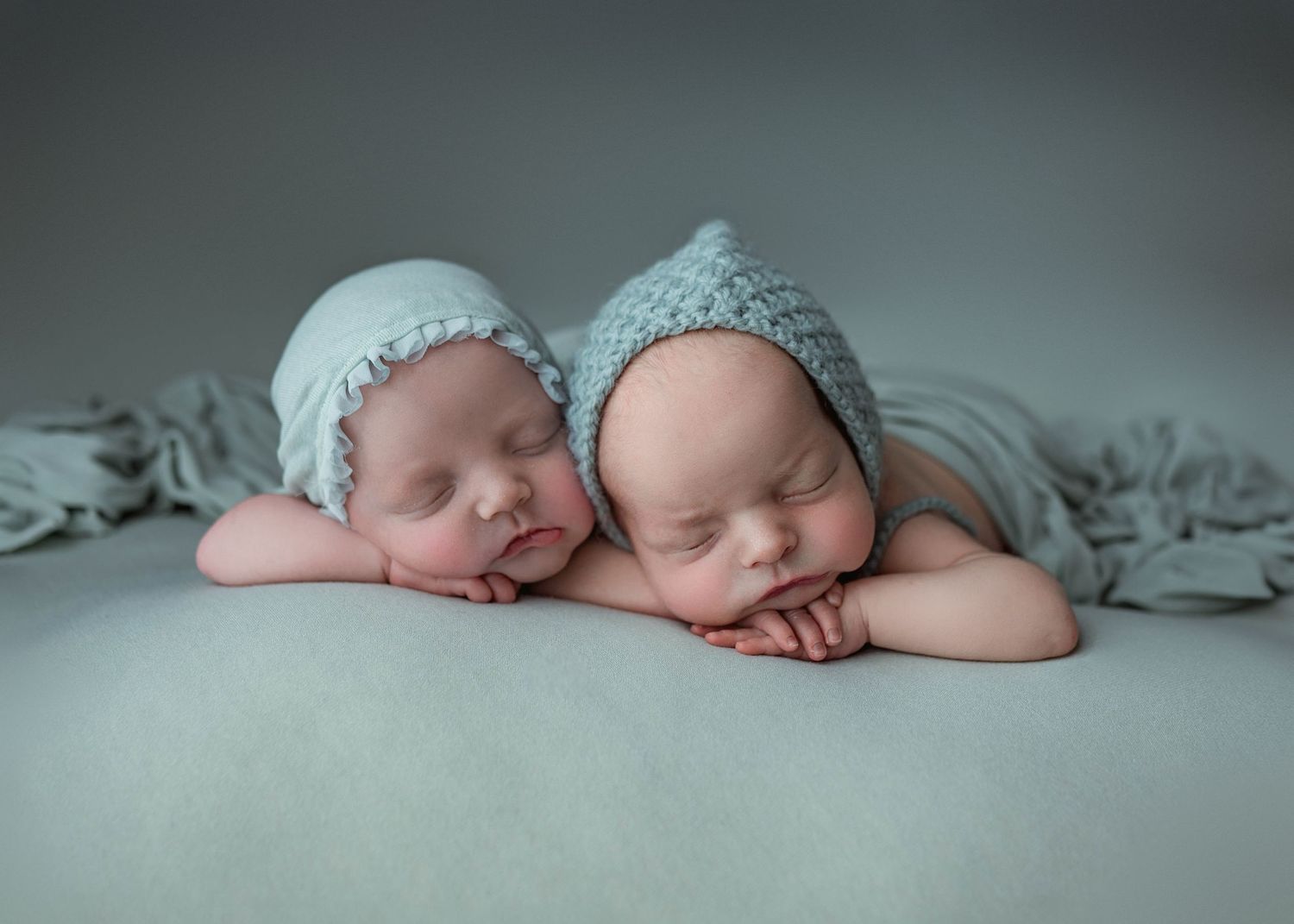 Twin newborn babies wearing matching blue knit bonnets sleep peacefully side by side on a gray backdrop.