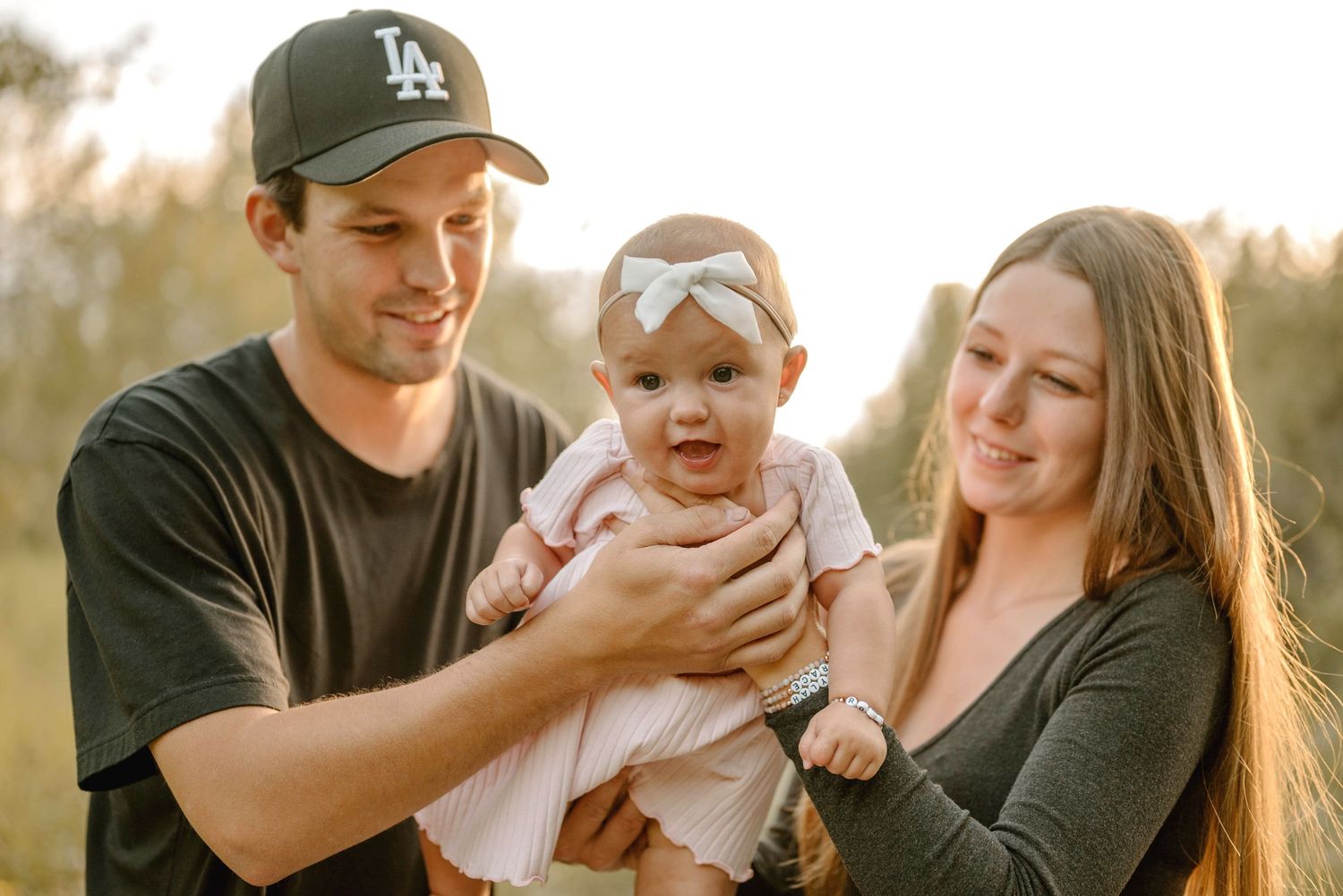 Young couple smiling and holding their baby girl outdoors in warm golden light.