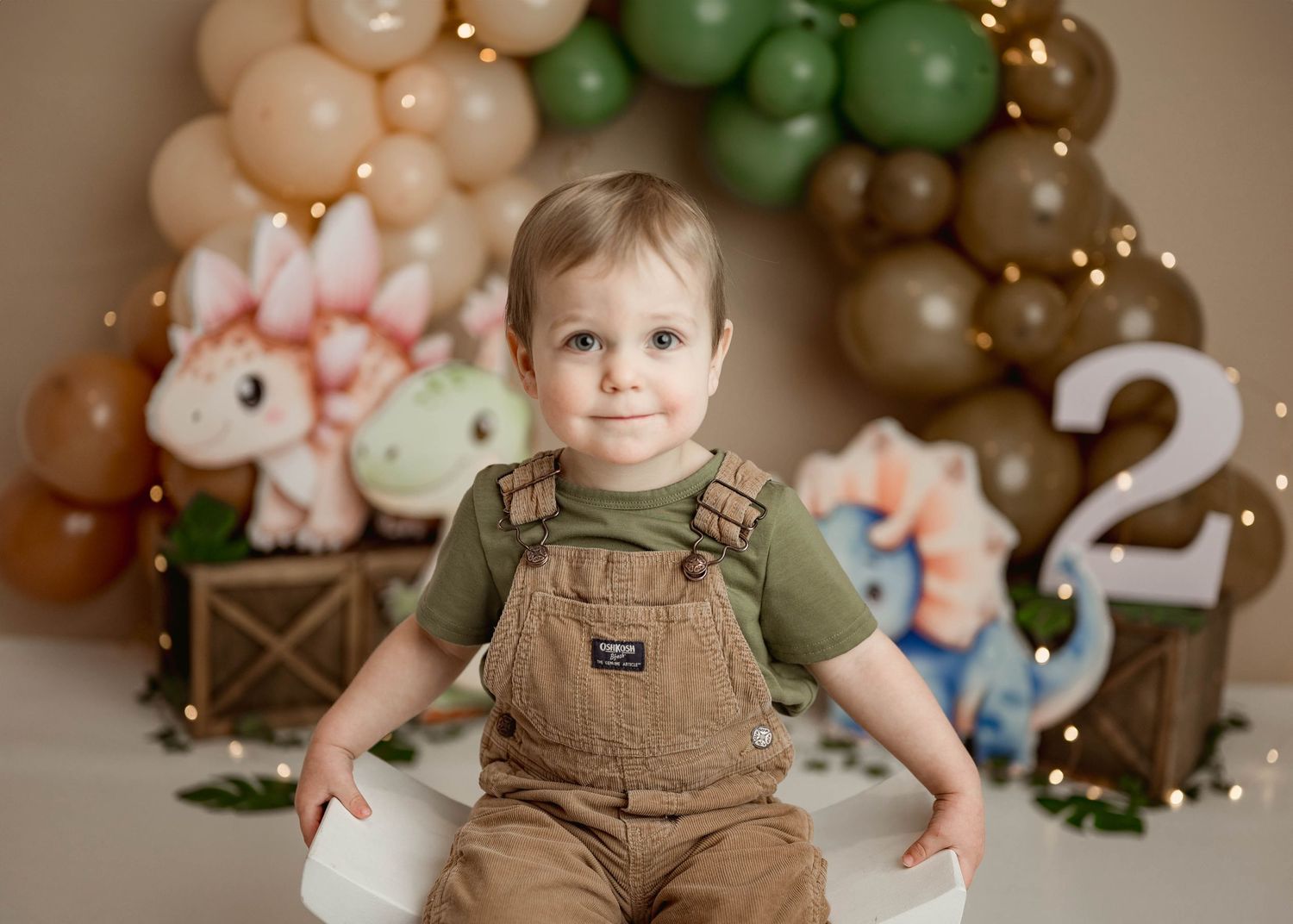 Toddler boy in brown overalls smiles at camera in front of a dinosaur themed second birthday balloon backdrop in Leduc.