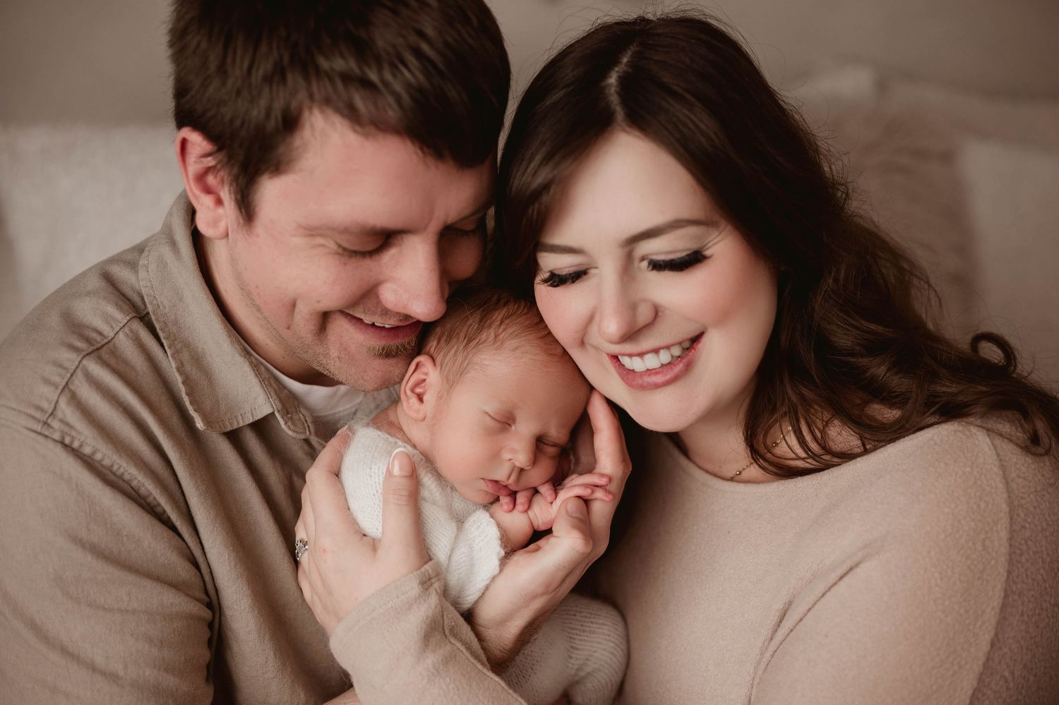 Happy couple smiling and cuddling their sleeping newborn baby in a warm cozy indoor setting.