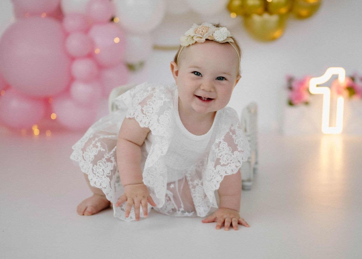 Smiling baby girl in white lace outfit crawls on floor at first birthday with pink and gold balloon backdrop.