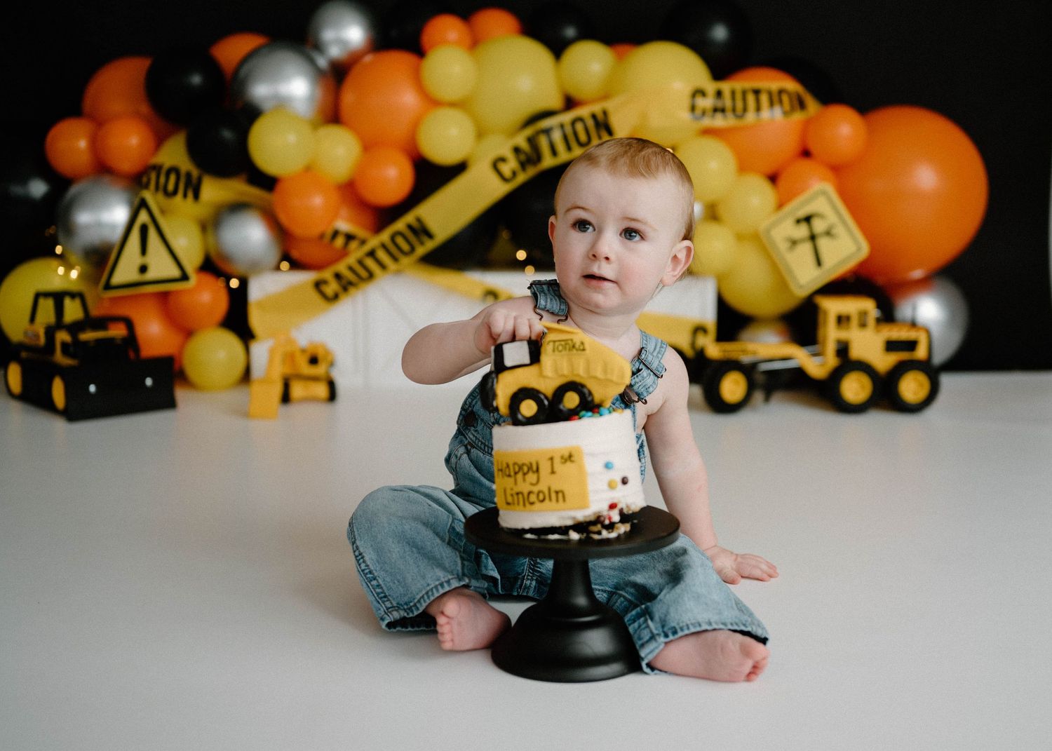 Baby boy in denim overalls reaches into construction-themed first birthday cake with toy dump truck topper.