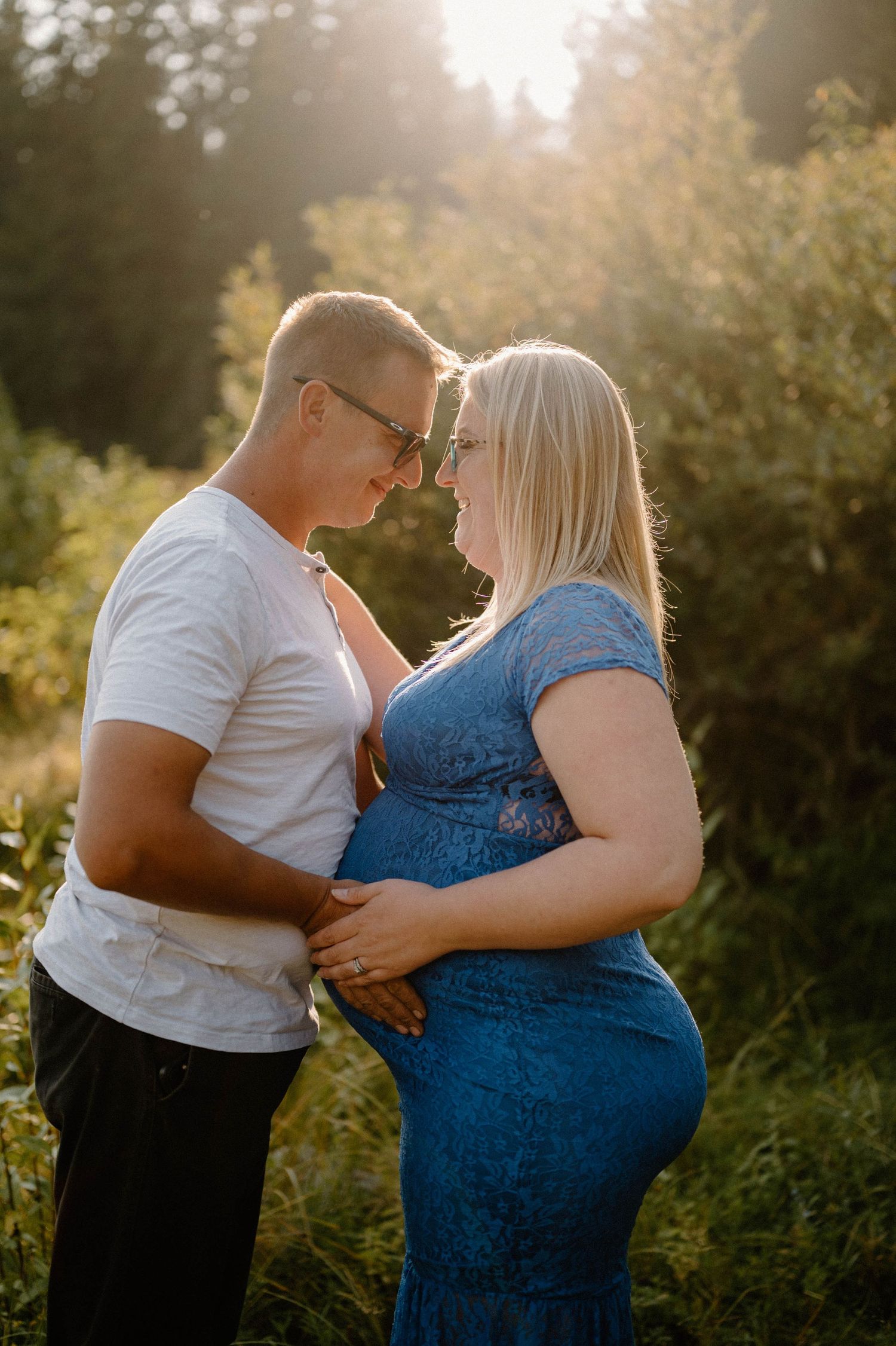 Expecting couple touching foreheads tenderly in golden sunlit forest setting.