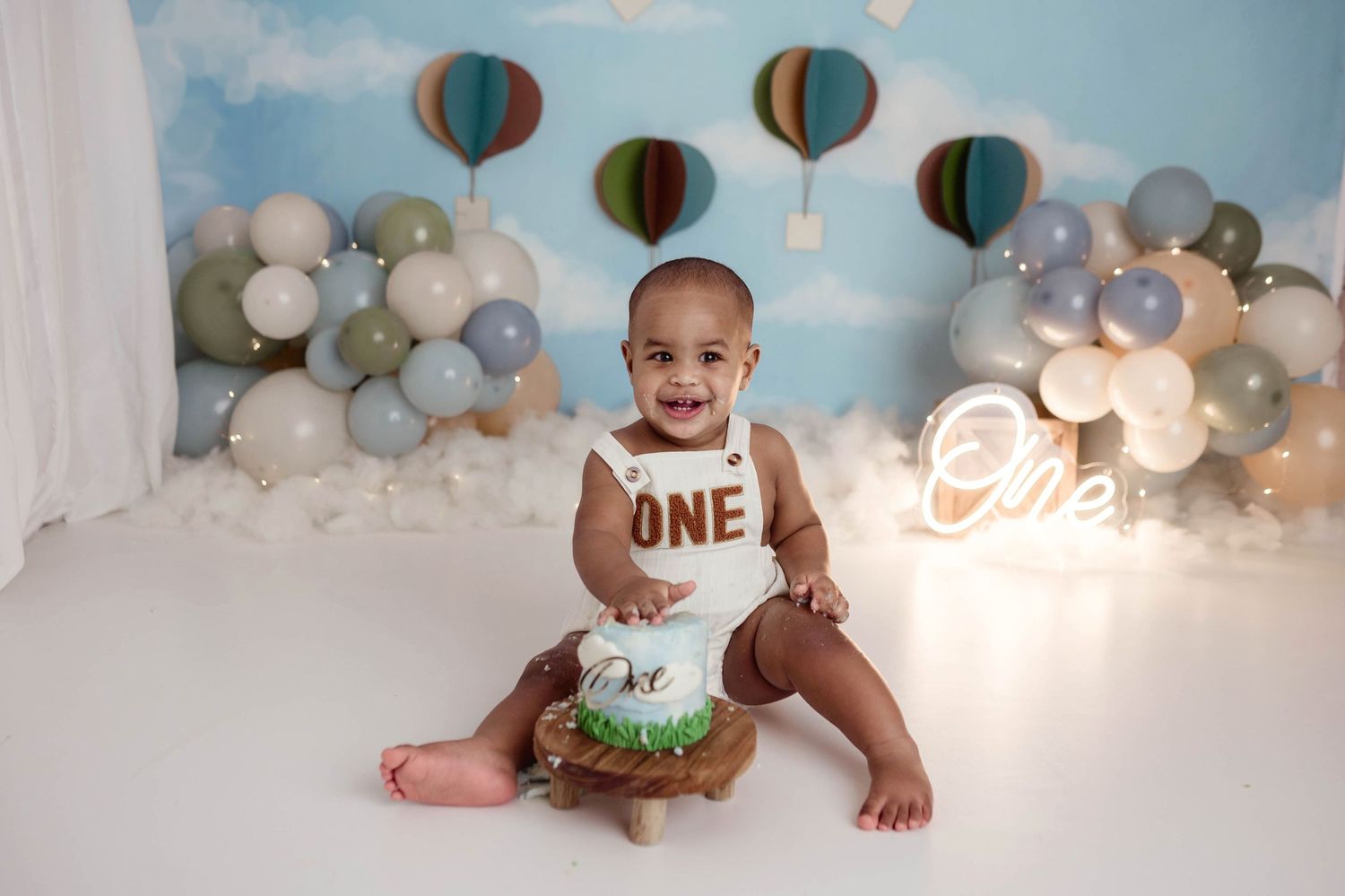 Baby boy in ONE romper smashing a cake at a hot air balloon themed first birthday session with balloon garland backdrop.