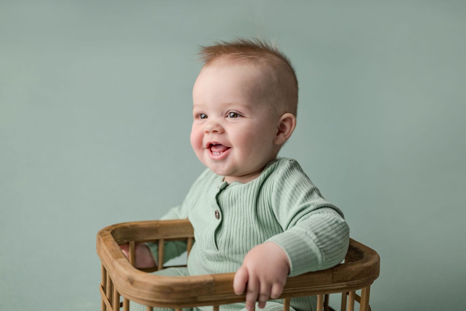 Cheerful baby boy in green ribbed onesie laughing while sitting in a wicker basket on teal.