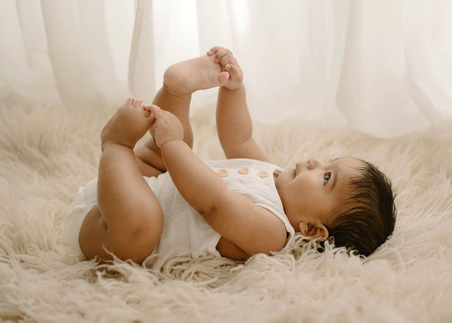 Playful baby in a white romper lying on a fluffy cream rug, holding their feet up in the air.