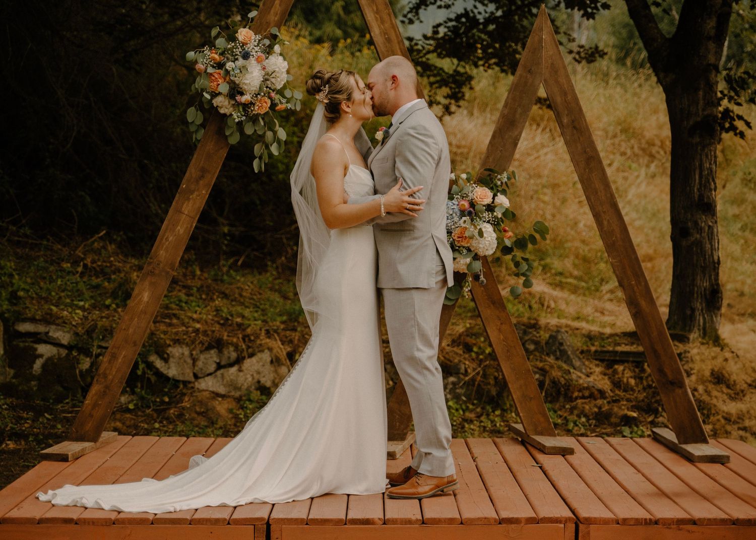 Bride and groom sharing a kiss under a rustic wooden triangular arch adorned with floral arrangements.