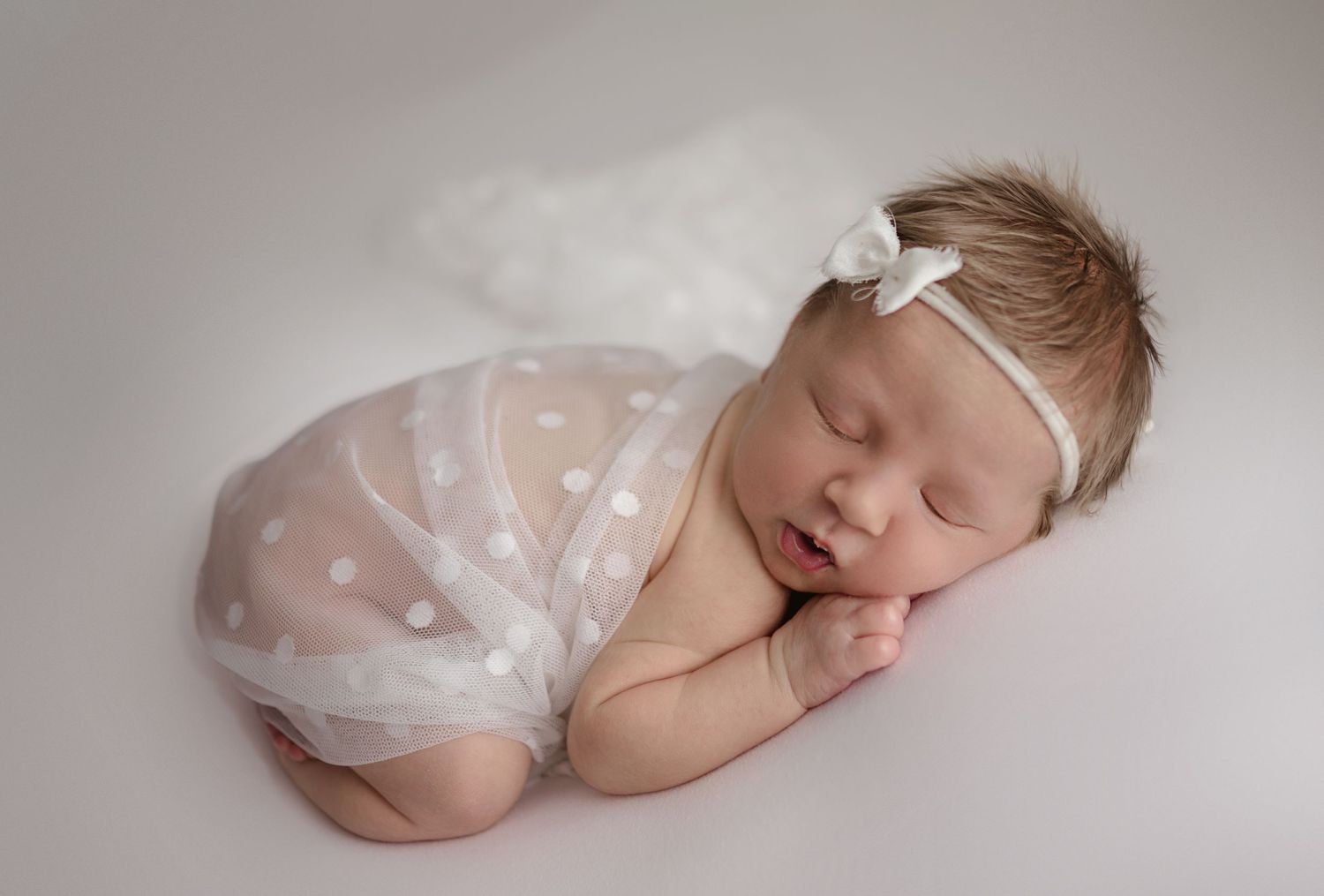 Sleeping newborn baby girl in white polka dot wrap and bow headband on soft white background.