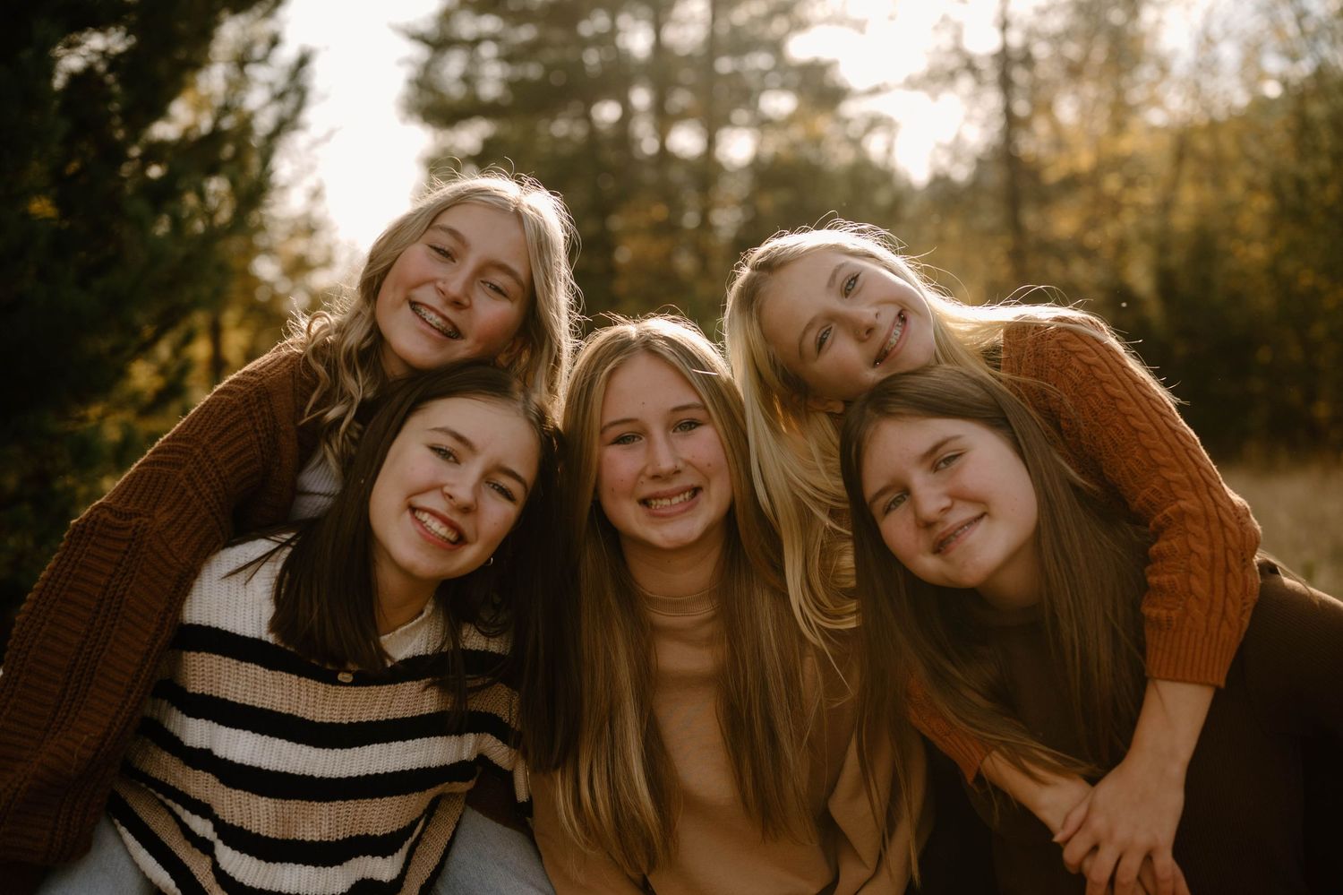 Five smiling teenage girls posing together outdoors in warm autumn tones surrounded by trees.