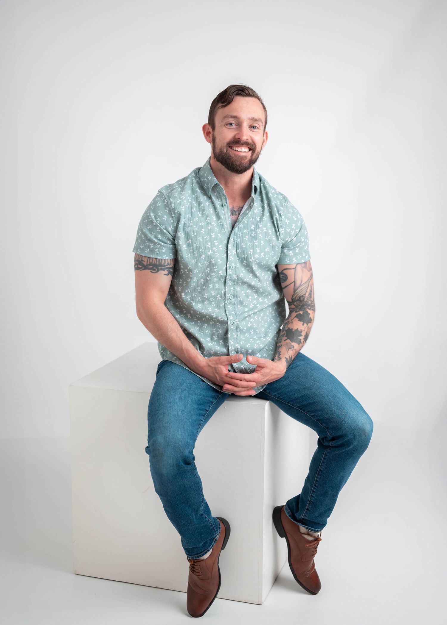 Smiling man with tattoos sits on white cube wearing teal patterned shirt and jeans in studio photo.
