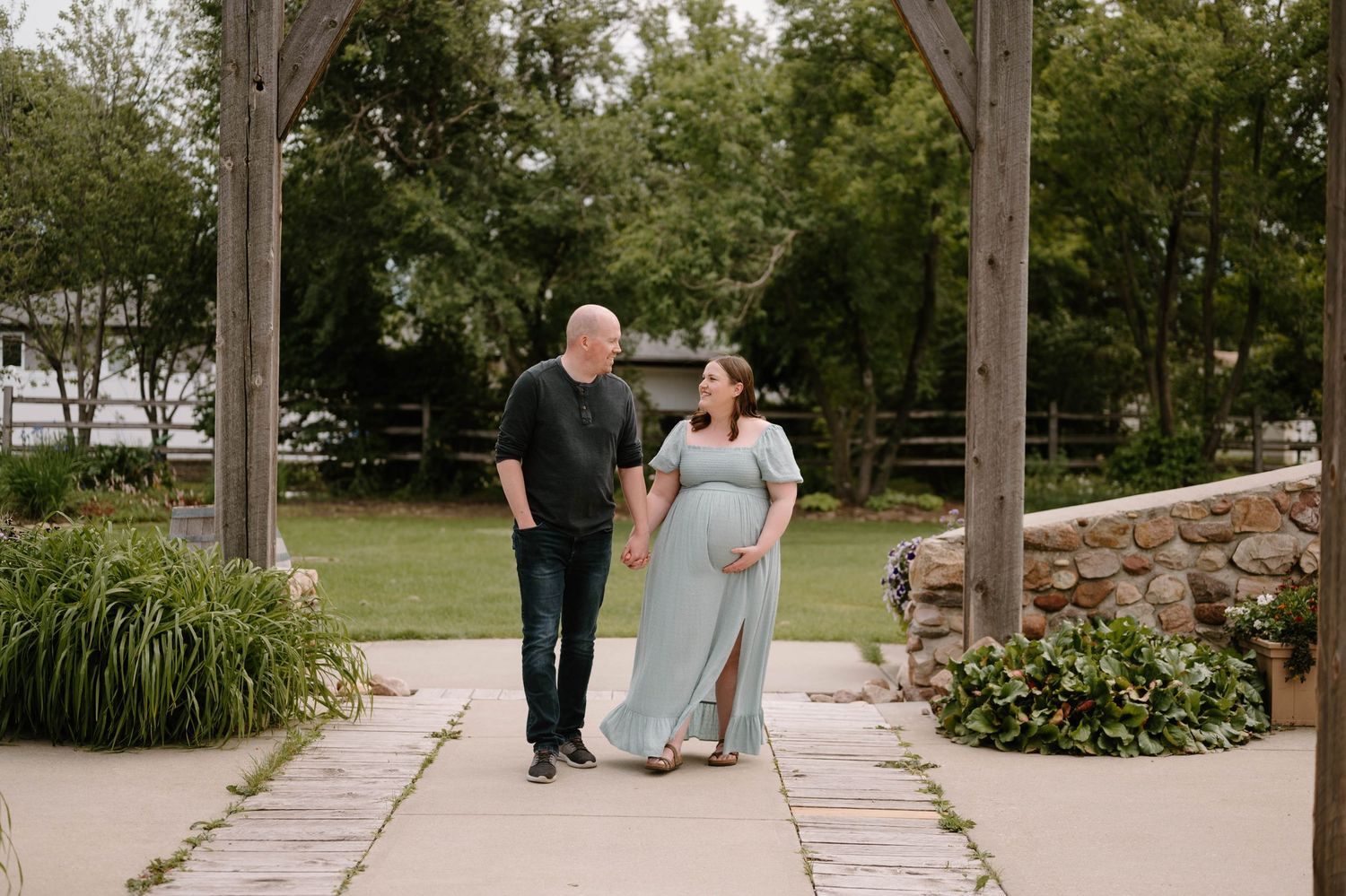 Pregnant couple walks hand-in-hand under wooden arbor in lush garden, woman in flowing sage green maternity dress.