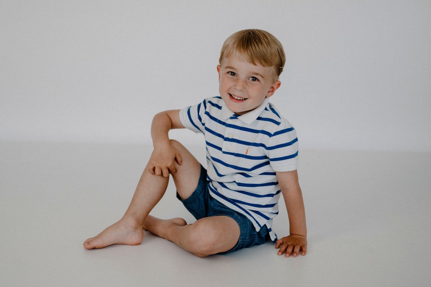 Smiling blonde boy in striped shirt and blue shorts sitting casually on white studio floor.