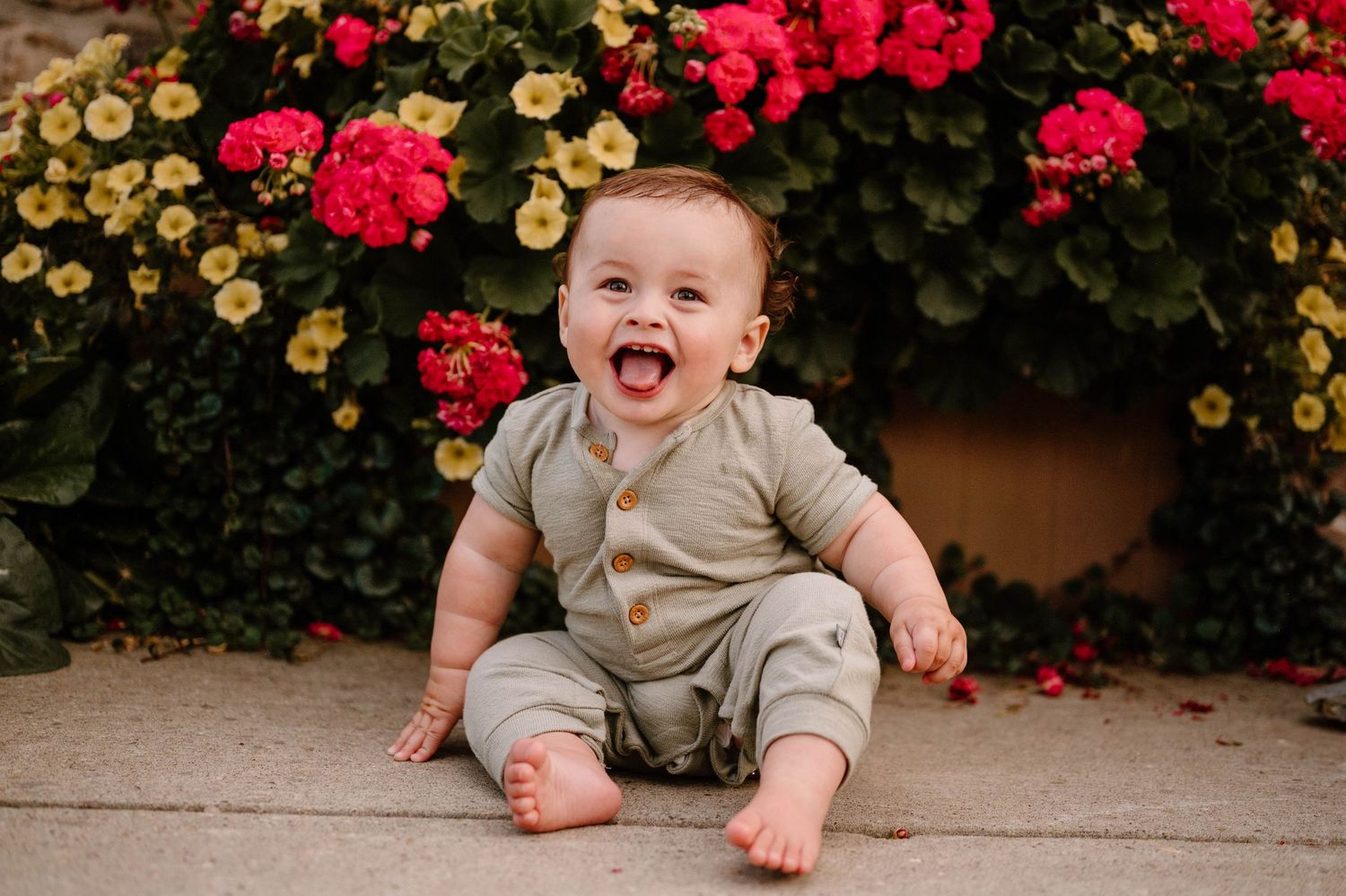 Joyful baby boy laughing while sitting barefoot in front of bright red and yellow garden flowers.