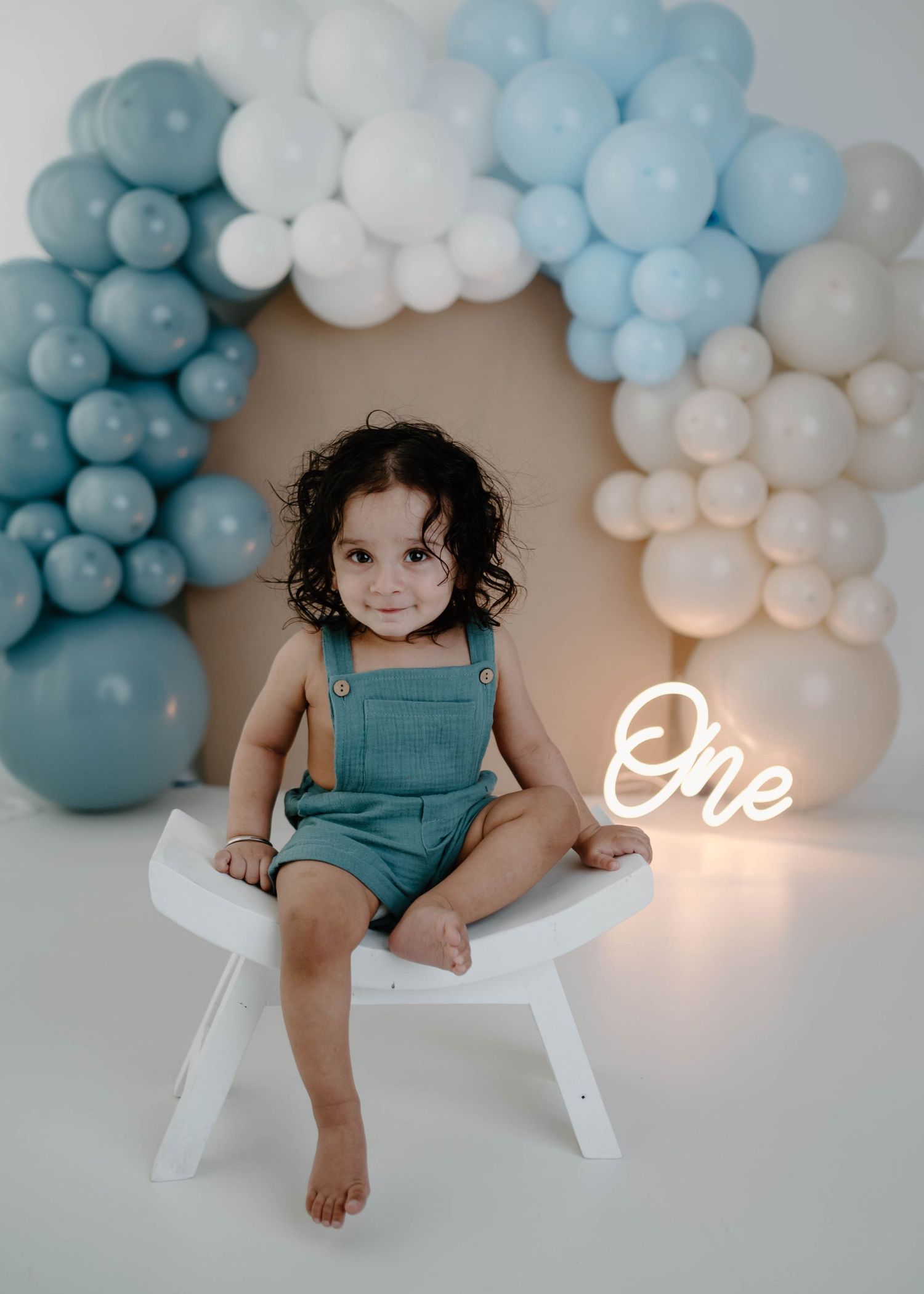 Smiling toddler in teal overalls sits on white stool with blue and white balloon arch and neon One sign.
