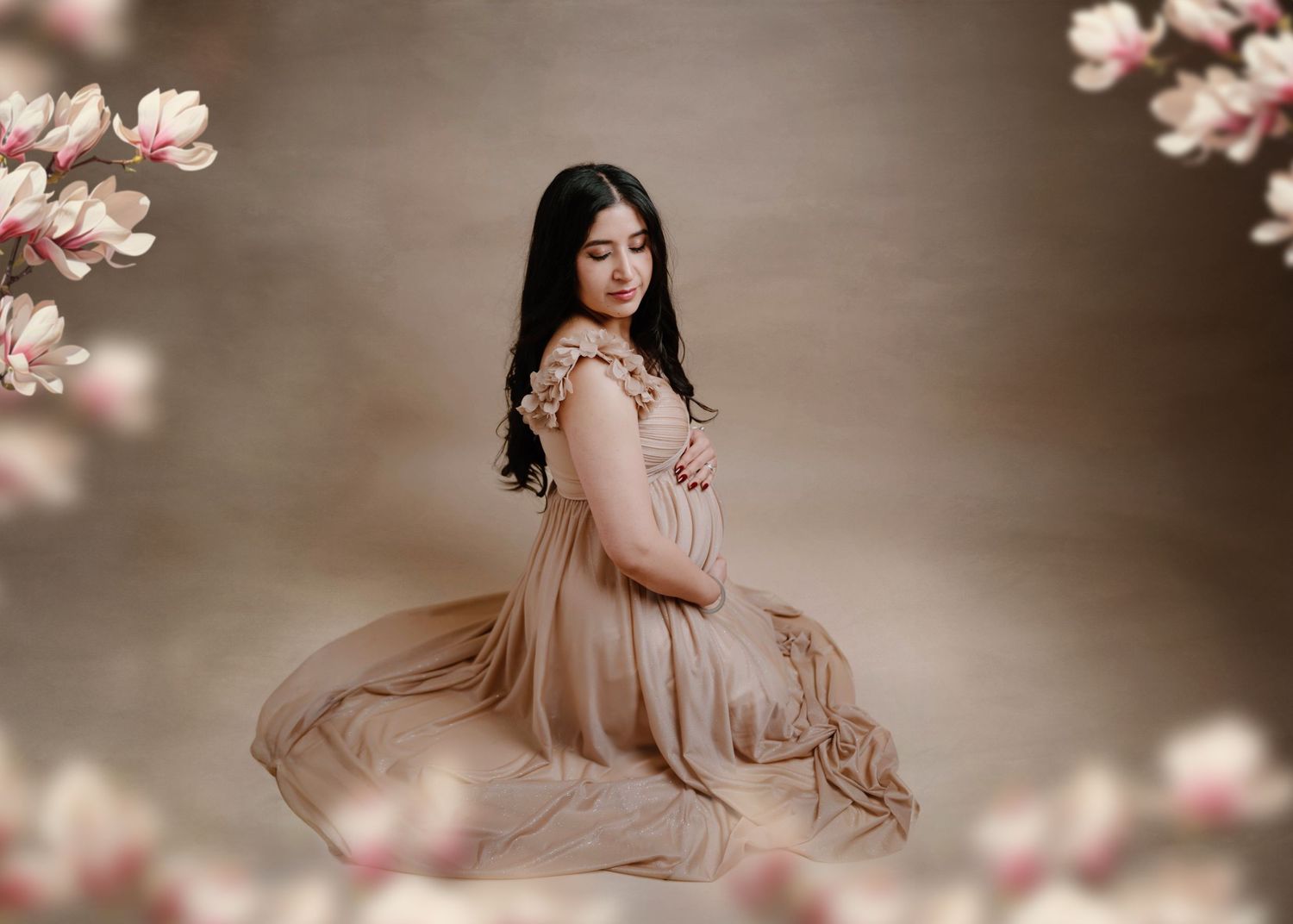 Expectant mother in flowing champagne dress kneeling gracefully surrounded by pink magnolia blossoms.