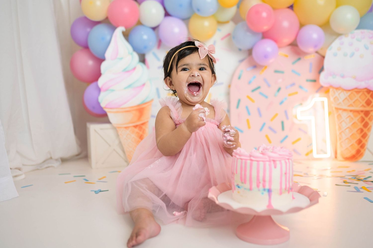 Excited baby girl in pink tutu smashes birthday cake at colorful ice cream themed first birthday party.
