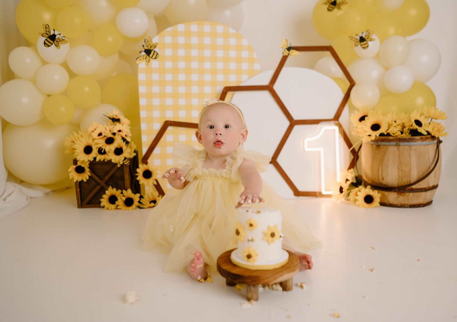 Baby girl in yellow dress smashes first birthday cake at bee-themed party with sunflowers and honeycomb decorations.