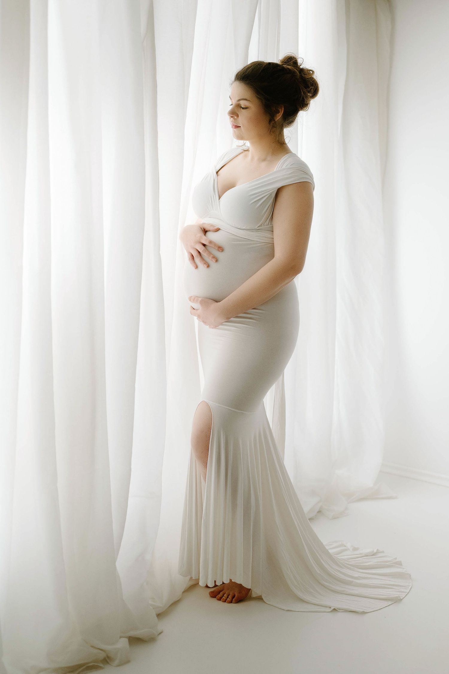 Elegant pregnant woman in white form-fitting mermaid gown stands between sheer white curtains in bright studio.