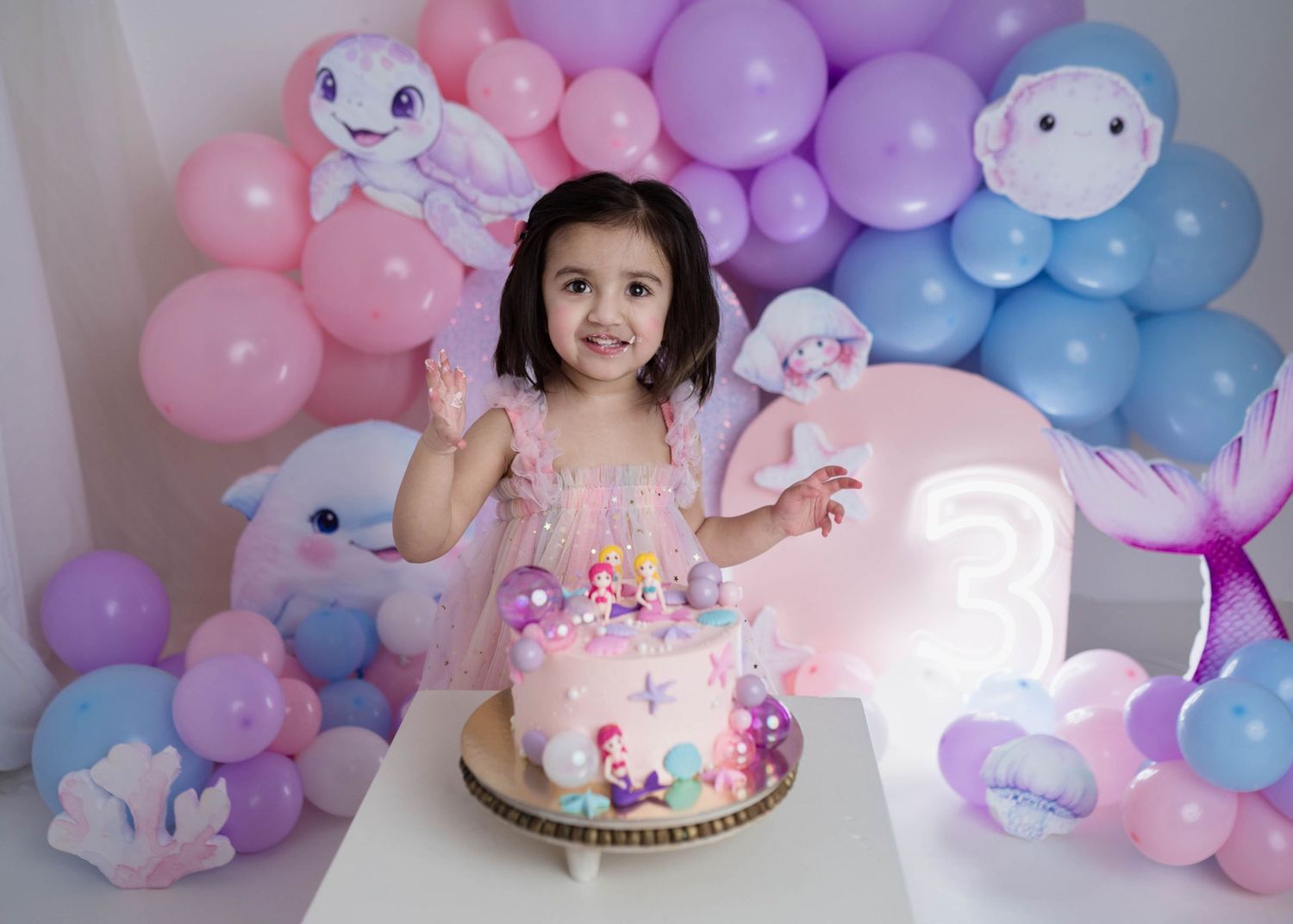 Cheerful toddler in pink dress celebrating third birthday with mermaid cake and balloon decorations.