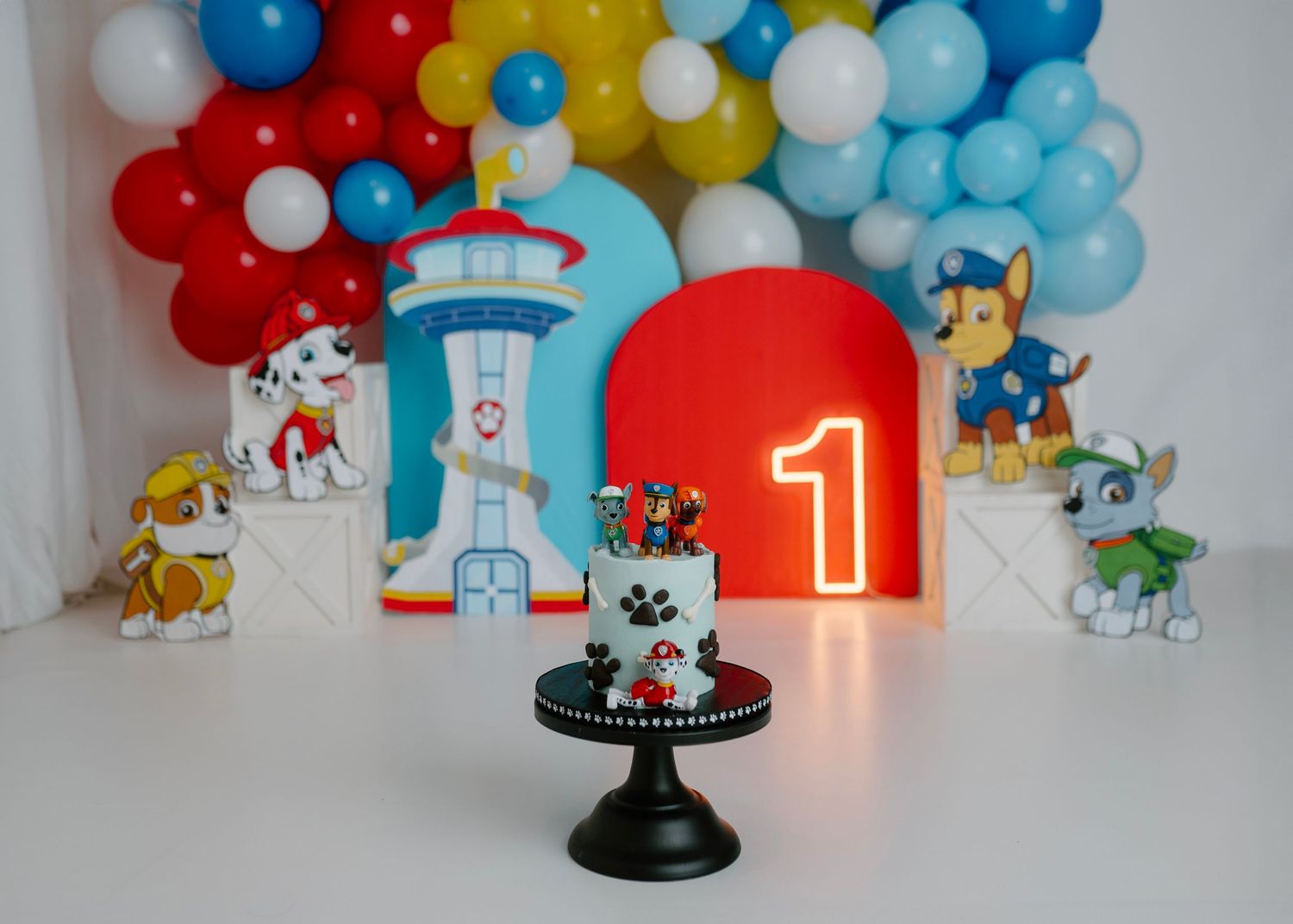Paw Patrol first birthday cake smash setup with character cutouts, balloon arch in red, blue, and yellow, and a number one neon sign.