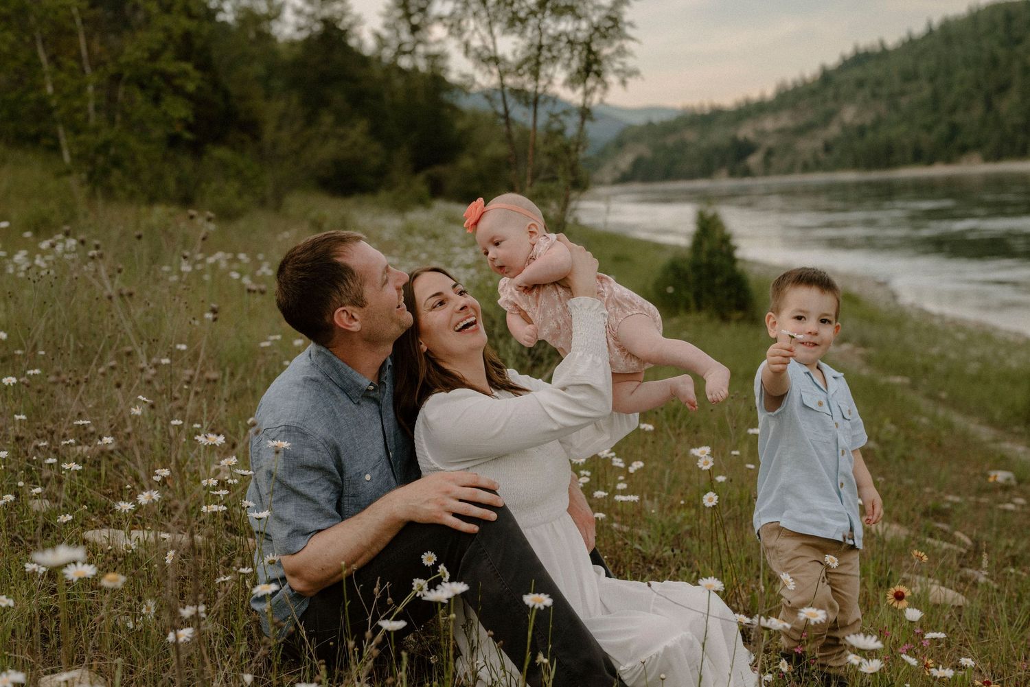 A joyful family sits in a wildflower meadow near a river at dusk.