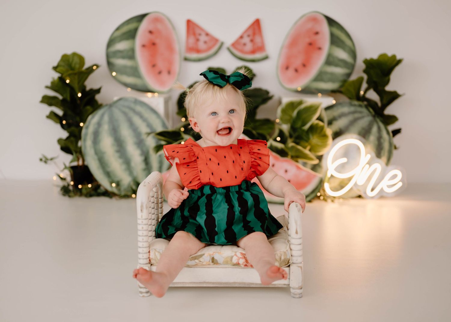 Baby girl in watermelon outfit sitting in a white chair surrounded by watermelon decorations for first birthday.