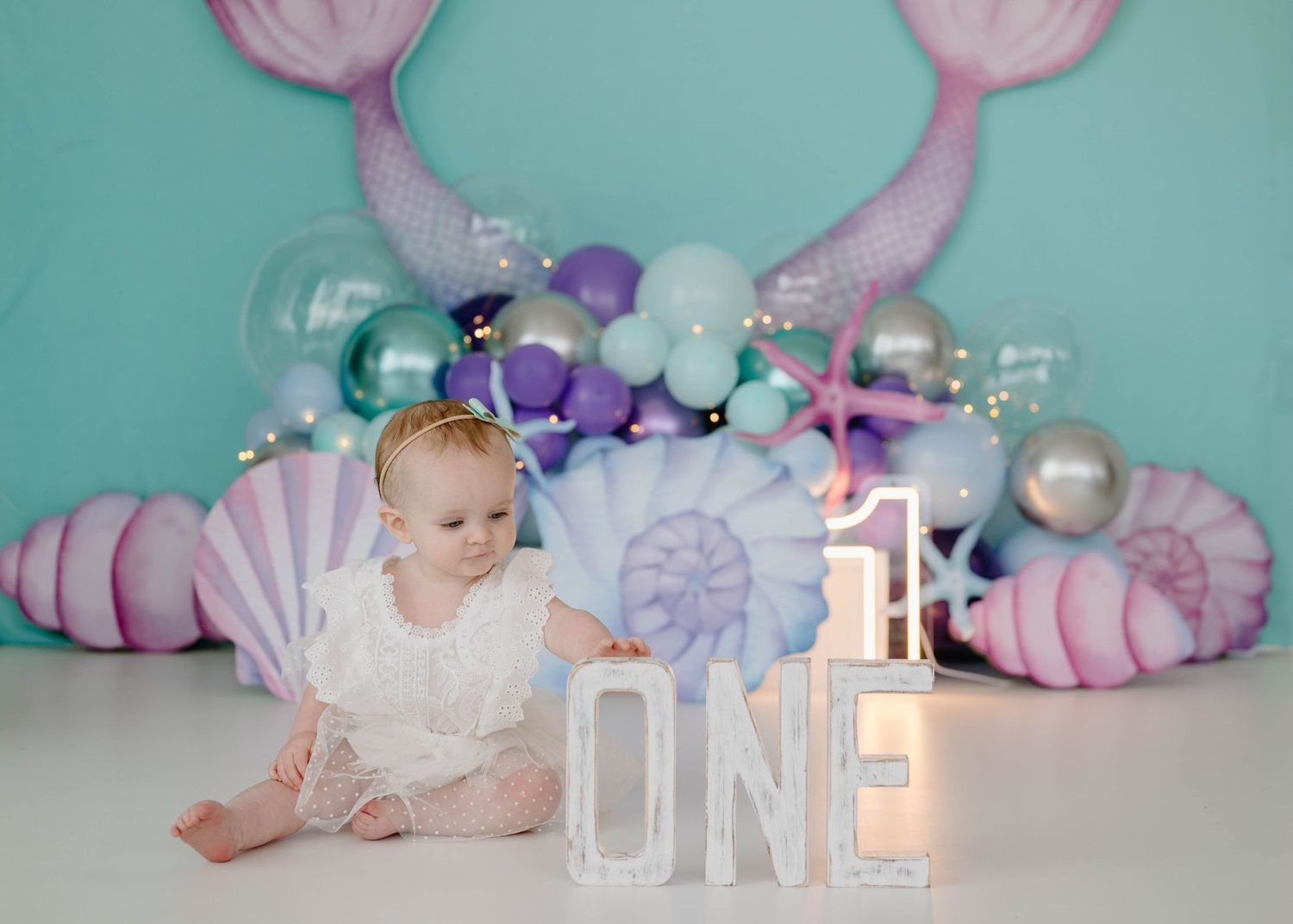 Baby girl in white dress sitting near ONE letters at mermaid-themed first birthday party.