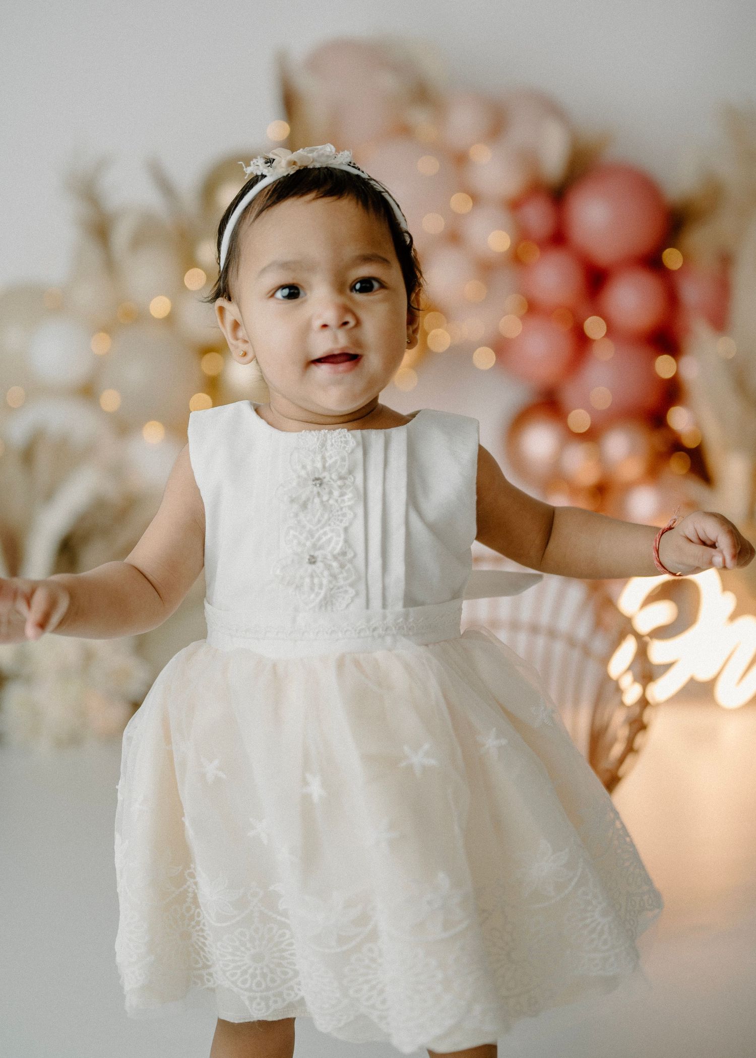 Adorable baby girl in white embroidered dress stands smiling at boho first birthday photo session.
