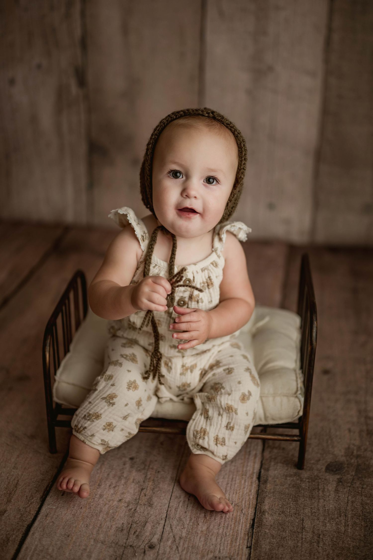 Sweet baby in floral romper and brown knit bonnet sits on miniature iron bed in studio portrait.