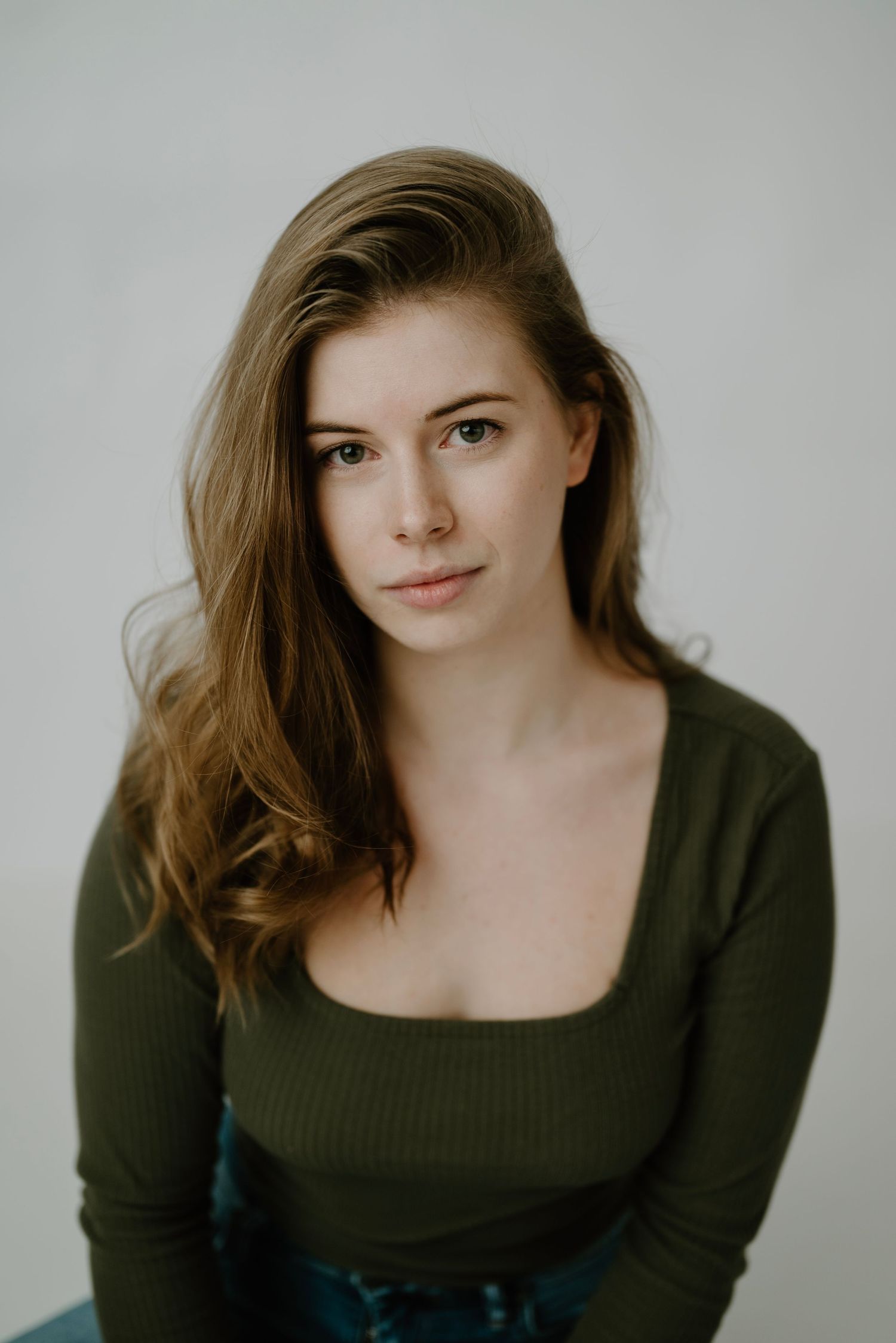 Young brunette woman in olive square-neck long-sleeve top with soft gaze on gray background.
