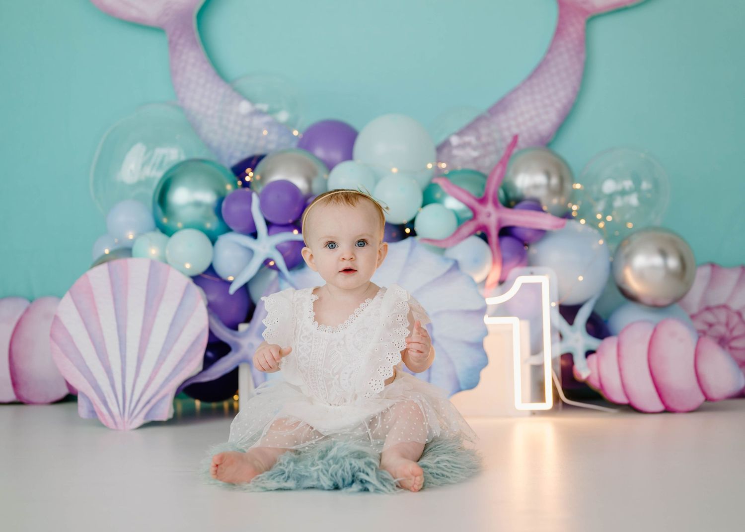 Baby girl in white dress at mermaid themed first birthday with shells and purple balloons.