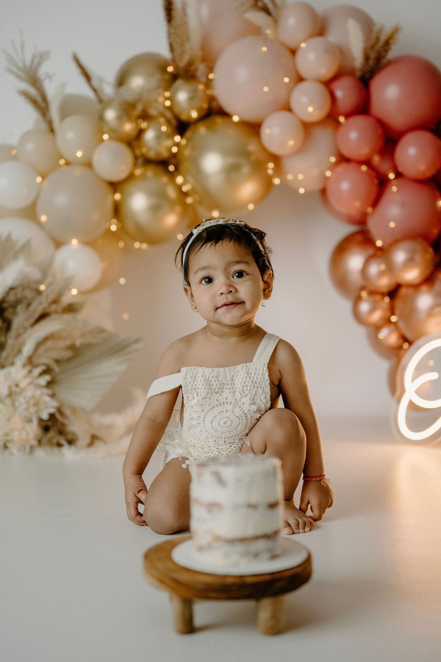 One year old baby girl in lace romper sits before smash cake with pink and gold balloon arch backdrop.