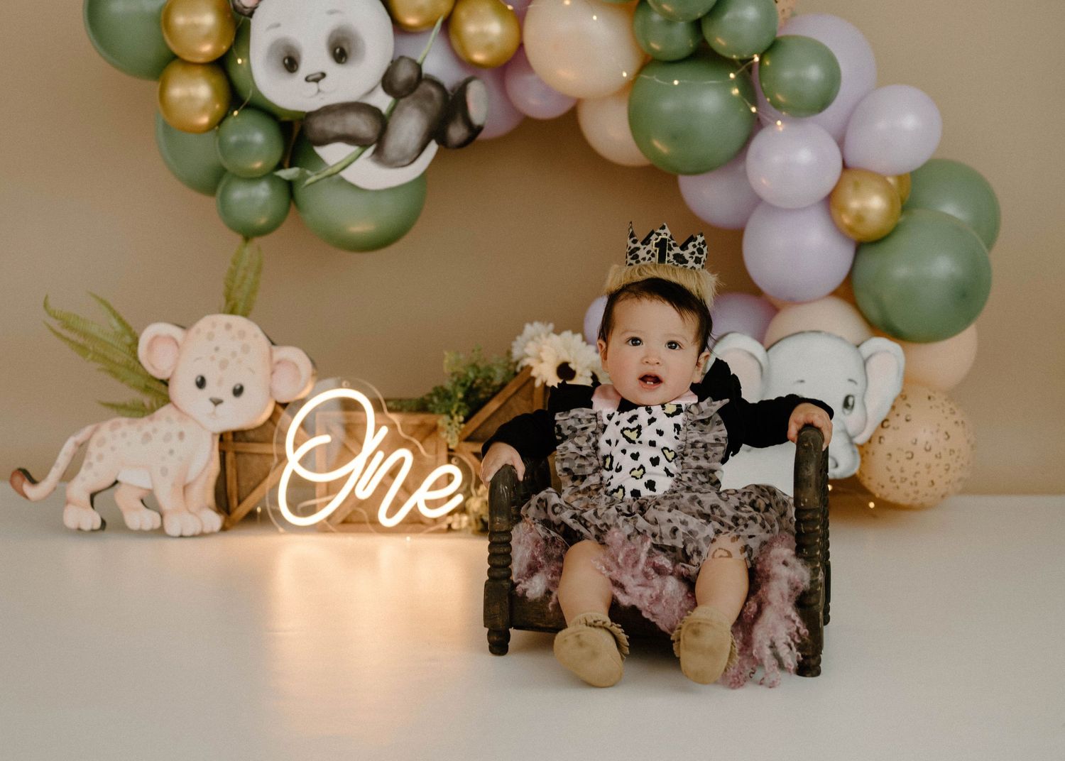 Baby girl in leopard outfit sits in chair beside ONE sign with jungle balloon arch.