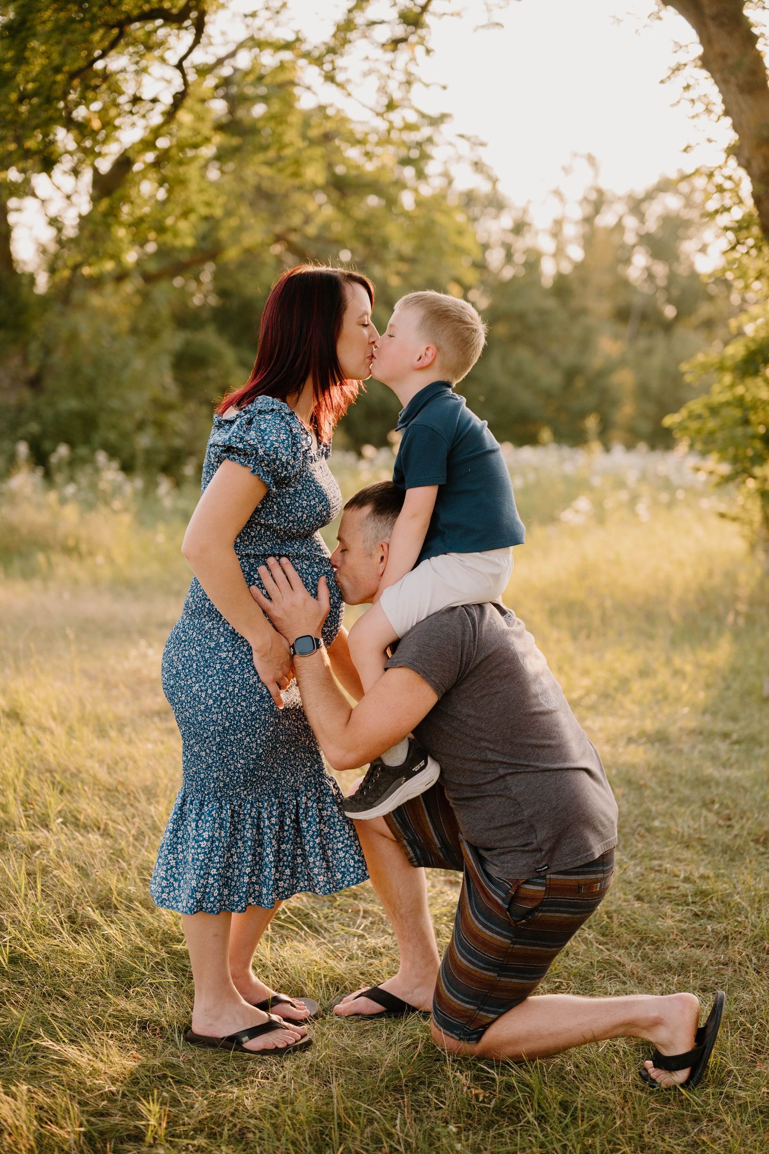 Pregnant woman kissing son while husband kneels kissing her baby bump in sunny park.